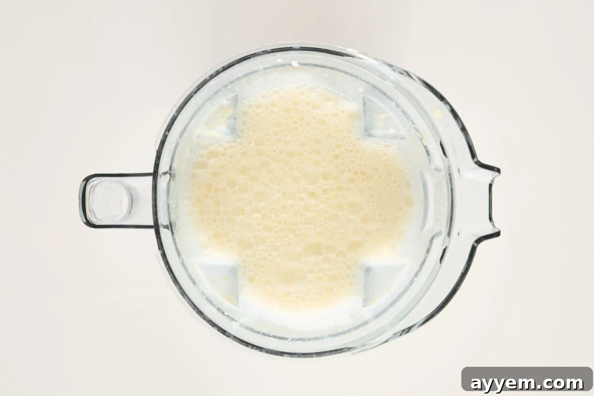 A top-down view of a blender filled with a light beige, frothy liquid, likely a smoothie or blended beverage, against a plain light background.