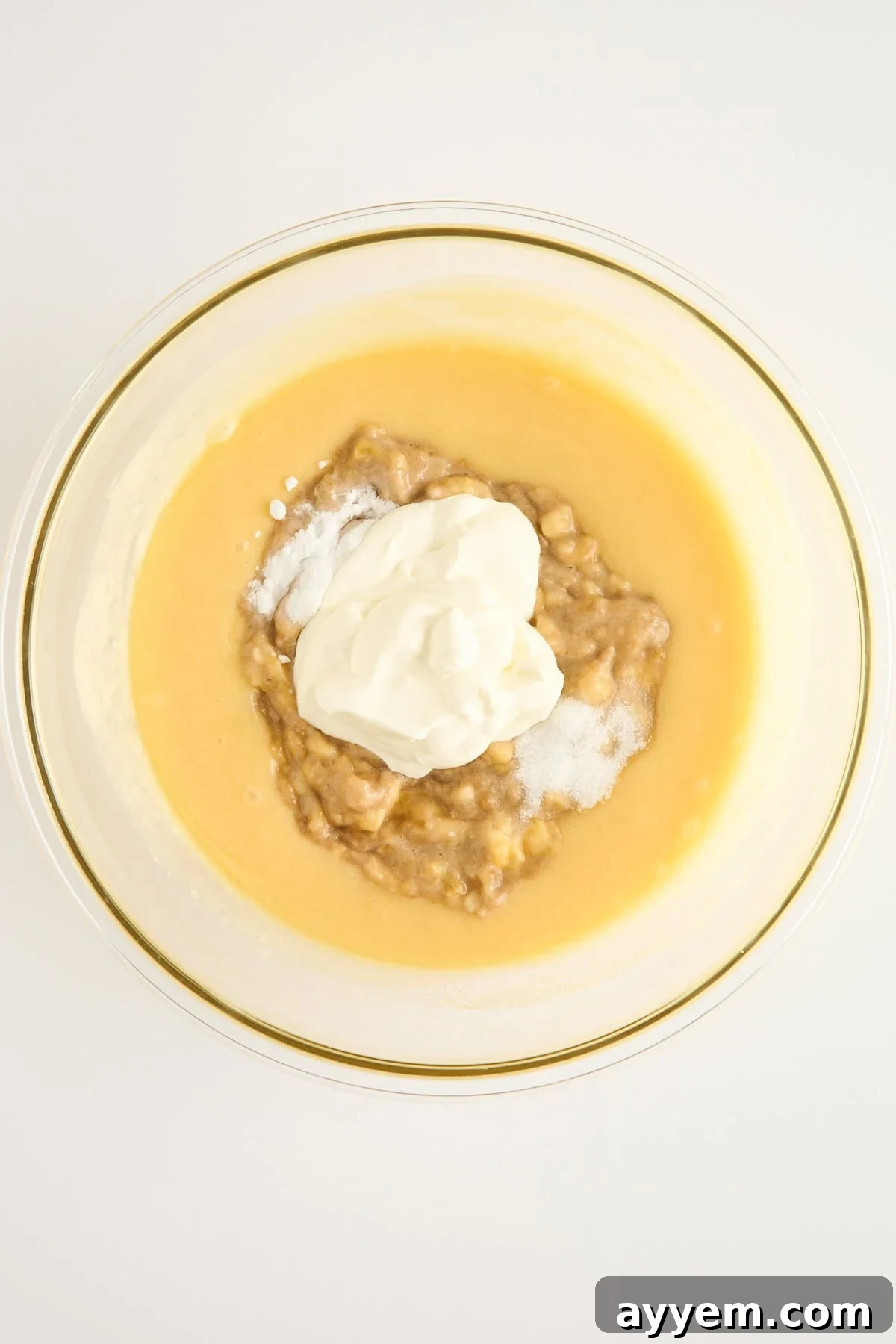 Mouthwatering Banana Blueberry Bread, Streusel Crowned 4 A glass bowl filled with a yellow batter, mashed bananas, white yogurt, and a sprinkle of baking soda, viewed from above on a white surface.