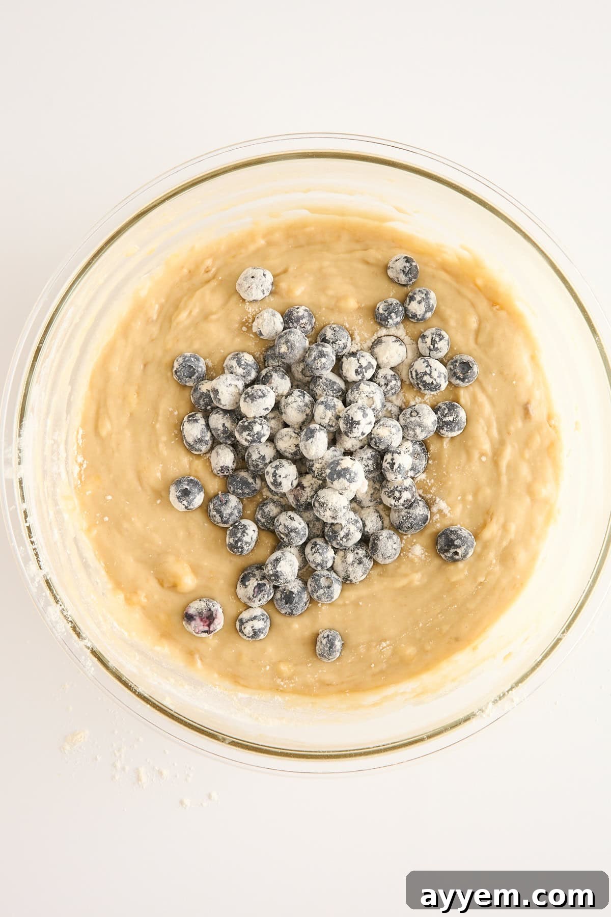 Mouthwatering Banana Blueberry Bread, Streusel Crowned 5 A glass bowl filled with muffin batter sits on a white surface. Fresh blueberries dusted with flour are piled on top, ready to be mixed into the batter. Some scattered flour is visible on the surface nearby.