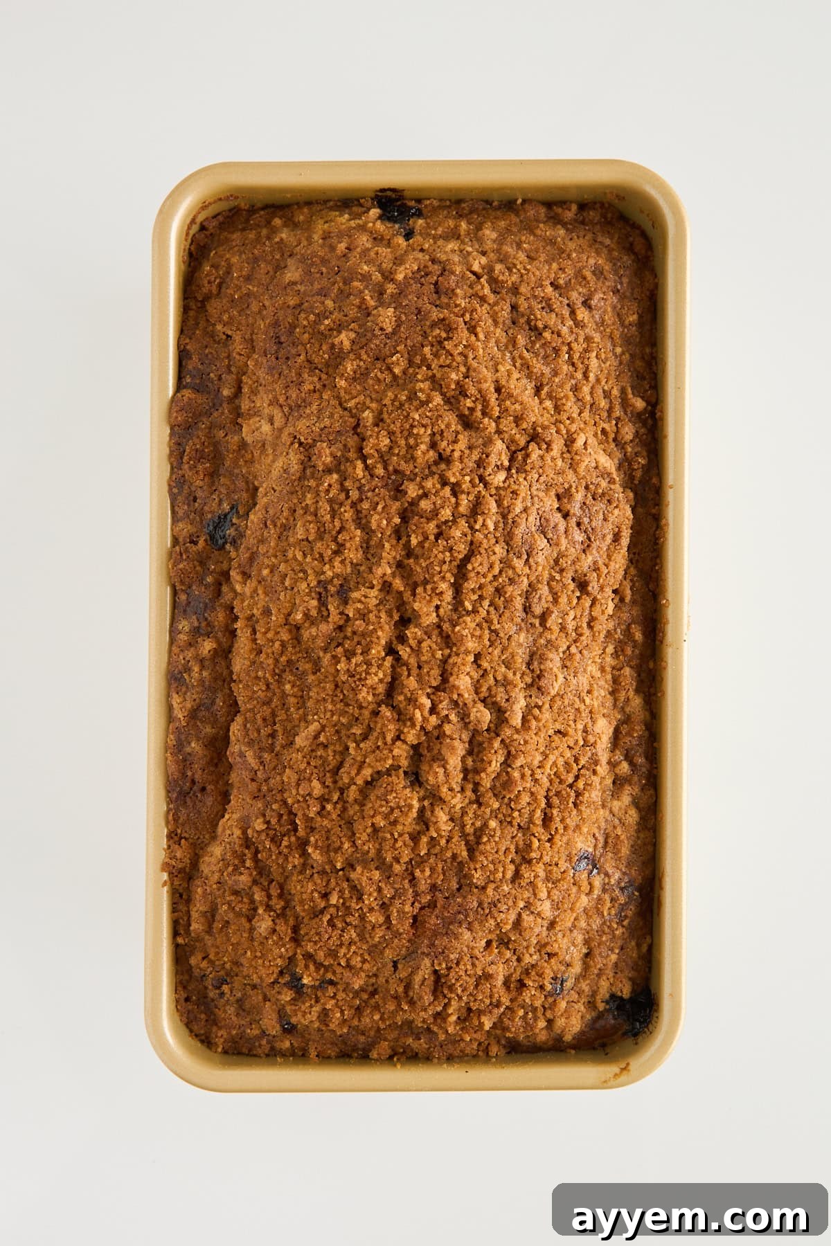 Mouthwatering Banana Blueberry Bread, Streusel Crowned 7 A rectangular loaf of baked bread with a crumbly brown crust is shown in a tan loaf pan, viewed from above on a white background.
