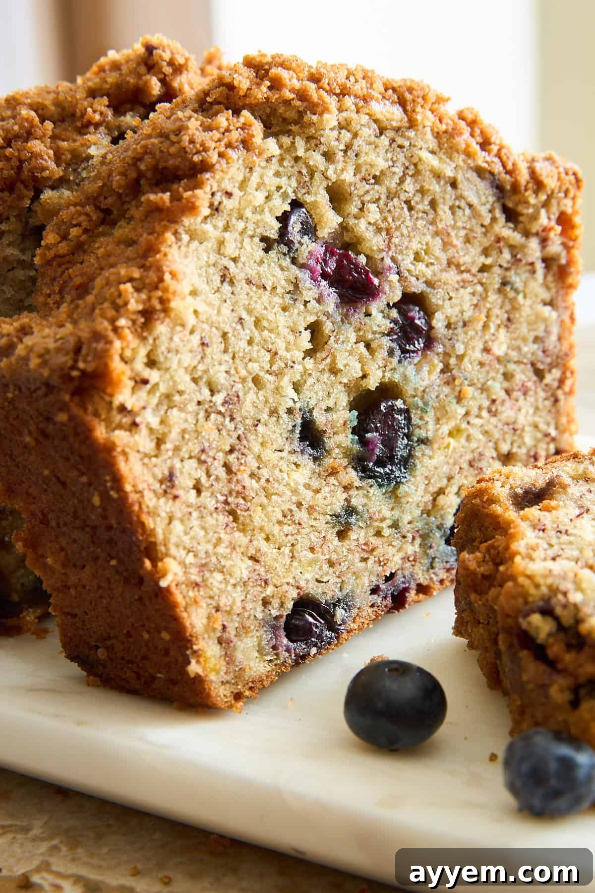Mouthwatering Banana Blueberry Bread, Streusel Crowned 8 A close-up of a sliced blueberry bread loaf with a crumbly topping, sitting on a white marble surface. Fresh blueberries are visible inside the bread and scattered nearby.