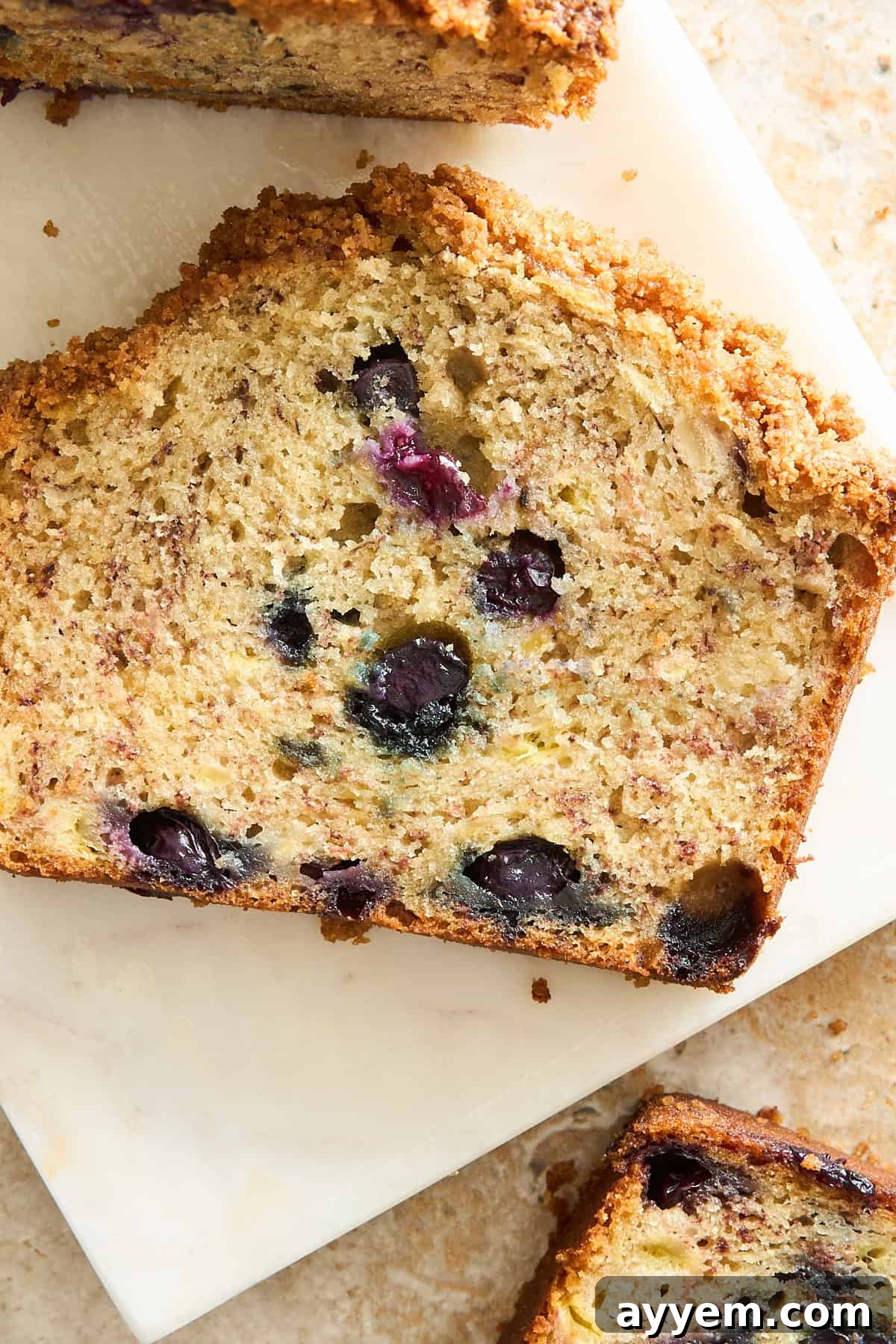 Mouthwatering Banana Blueberry Bread, Streusel Crowned 9 A close-up of a slice of blueberry zucchini bread on a white surface, showing a golden-brown crust and moist interior with visible blueberries and shredded zucchini.