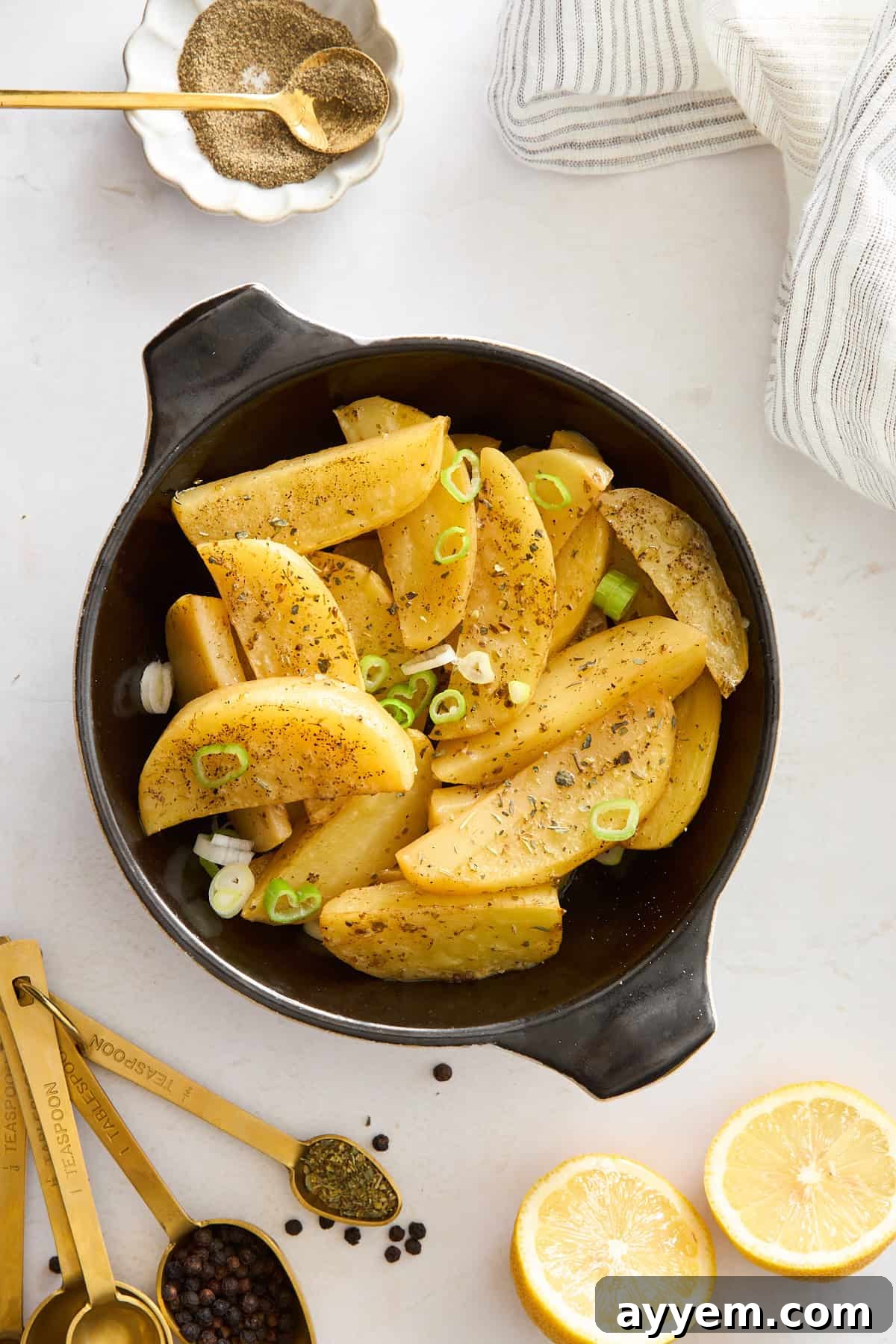A black dish filled with seasoned potato wedges garnished with sliced green onions, surrounded by measuring spoons with spices, lemon halves, and a striped napkin on a white surface.