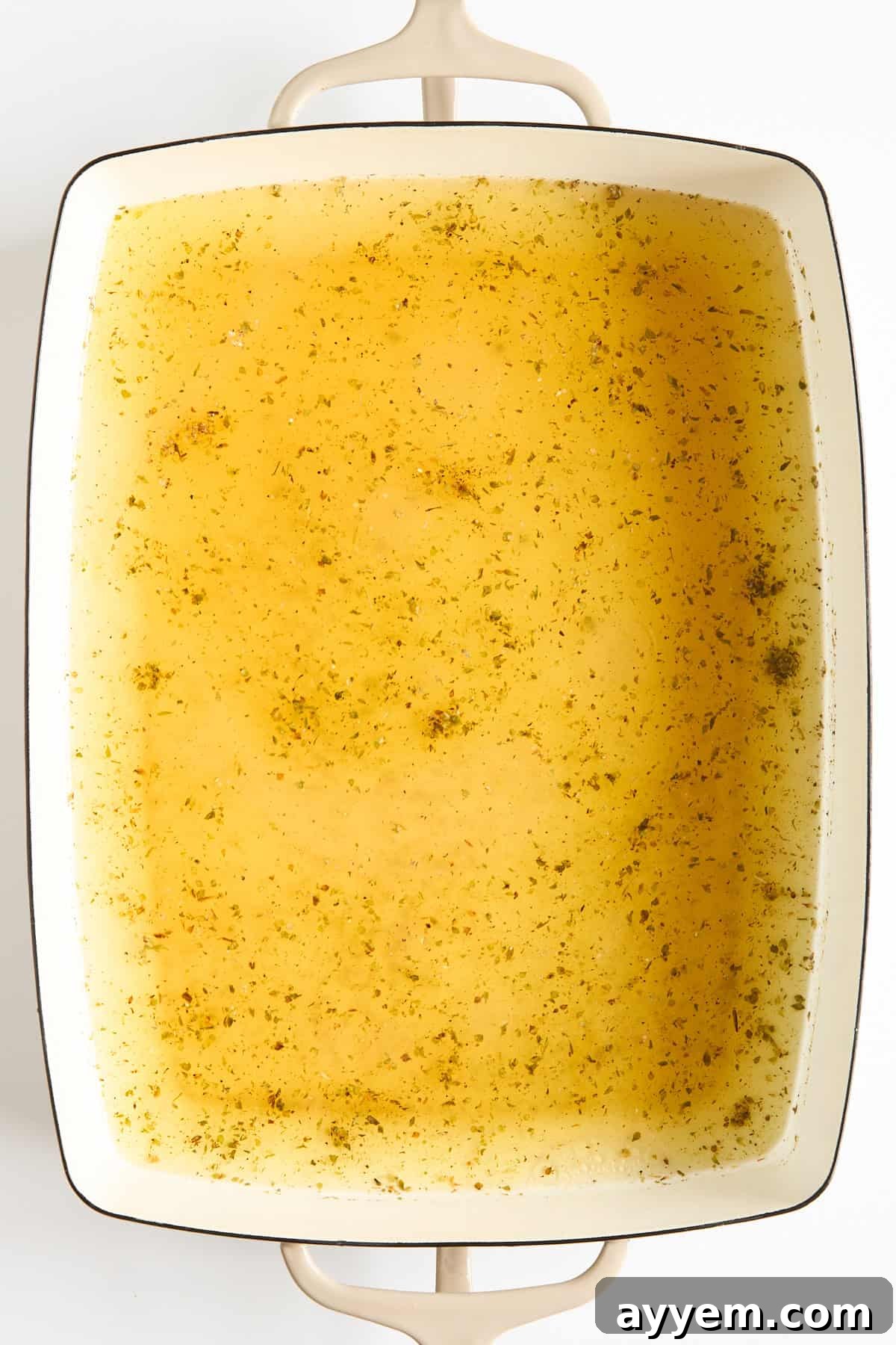 A rectangular white baking dish filled with a golden-yellow broth sprinkled with herbs, viewed from above. The dish has two handles and sits on a white surface.