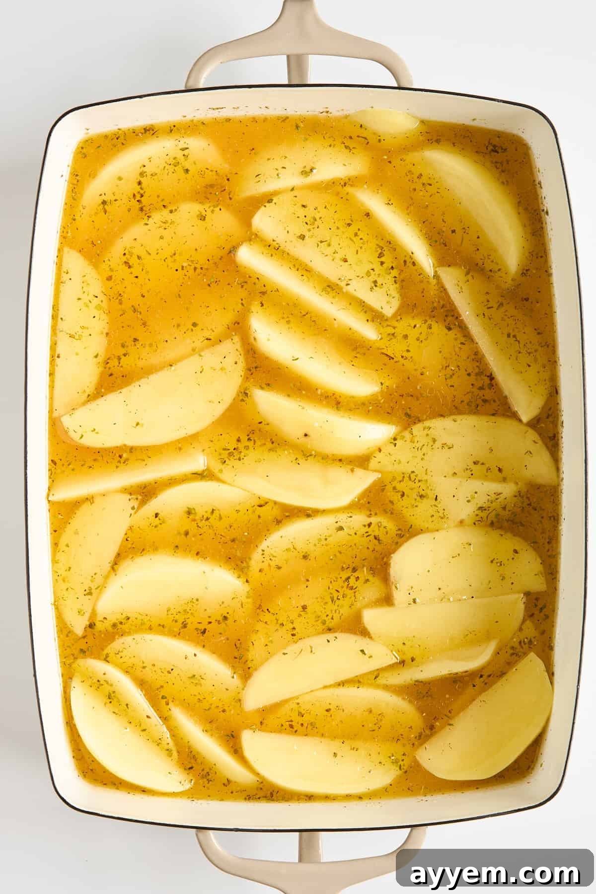 Sliced potatoes arranged in a white baking dish, covered in a yellow seasoned broth, ready to be cooked.