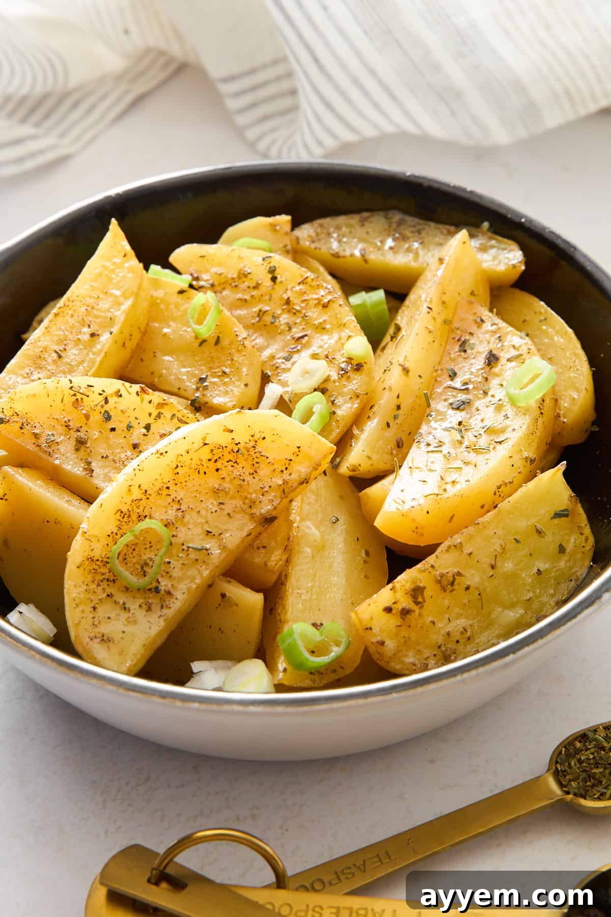 A bowl filled with seasoned potato wedges, garnished with chopped green onions and herbs, sits on a white surface next to golden measuring spoons and a striped cloth.