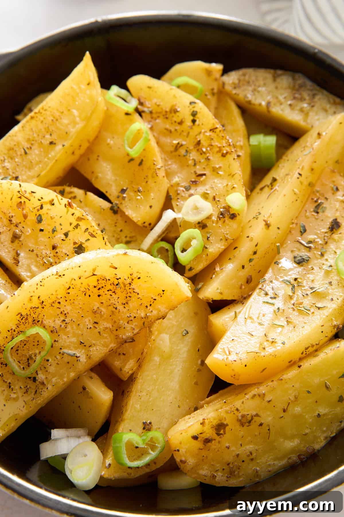 A bowl of seasoned potato wedges garnished with sliced green onions and herbs, showing a close-up of the golden, baked potatoes with visible spices on their surface.