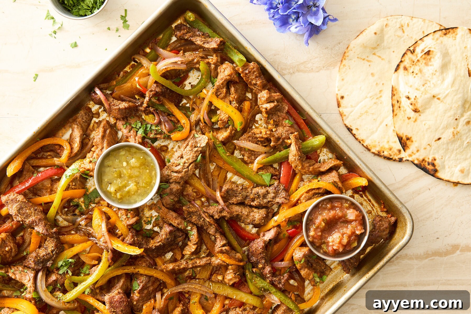 Sheet pan steak fajitas on a baking sheet with salsa verde and red salsa next to two tortillas. 