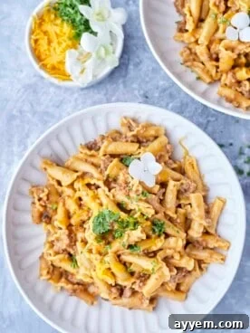 Two plates of one-pot cheeseburger pasta with a side bowl of shredded cheese.