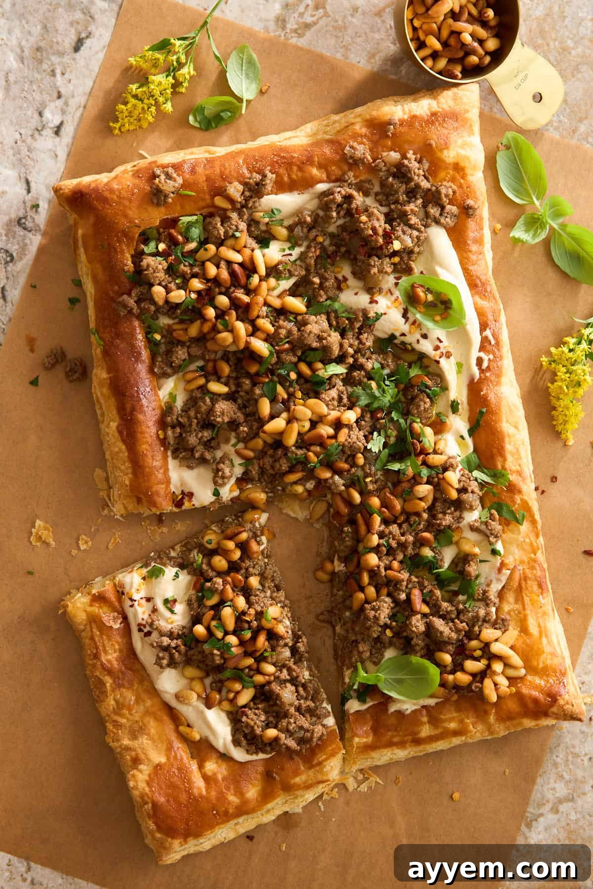 A rectangular puff pastry topped with ground meat, pine nuts, fresh herbs, and a creamy white sauce, partially cut into pieces and arranged on brown parchment paper with scattered yellow flowers and basil leaves nearby.