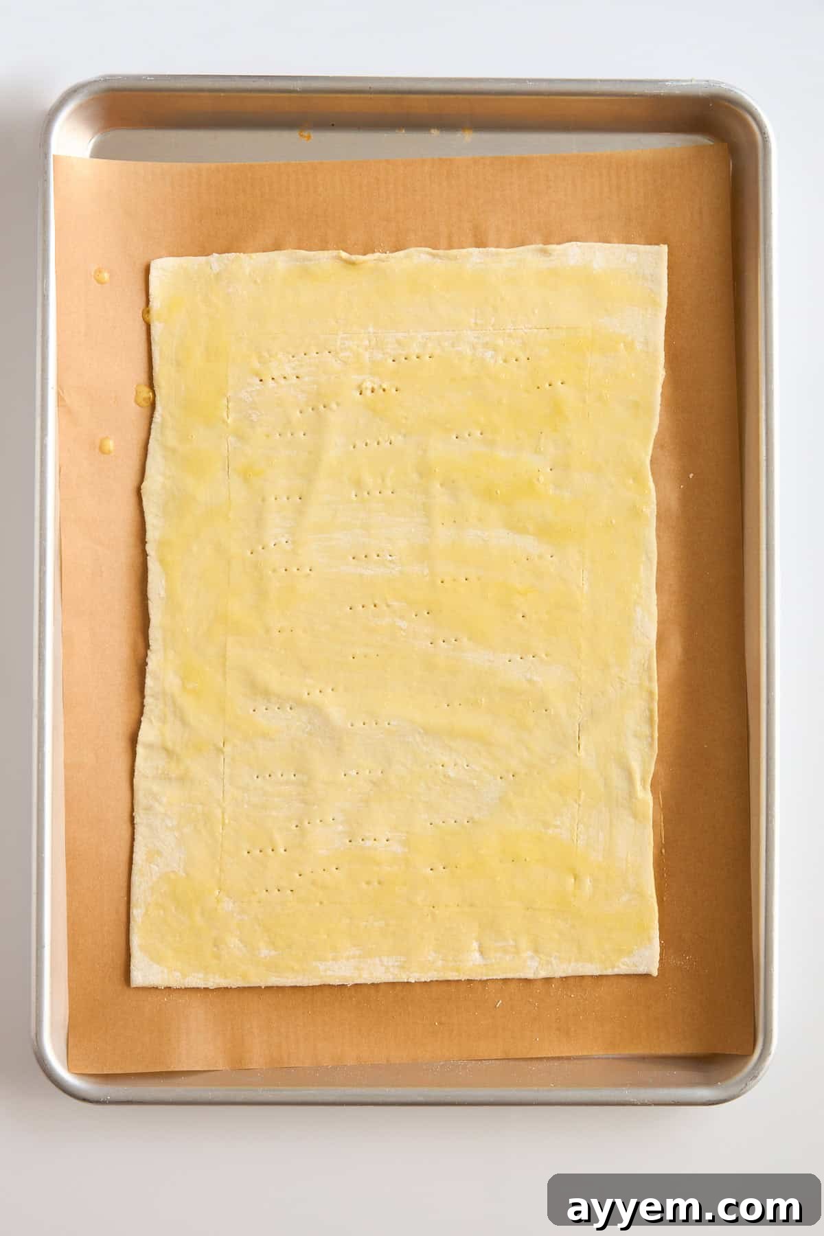A rectangular sheet of dough brushed with egg wash and docked with fork marks sits on parchment paper on a baking tray.