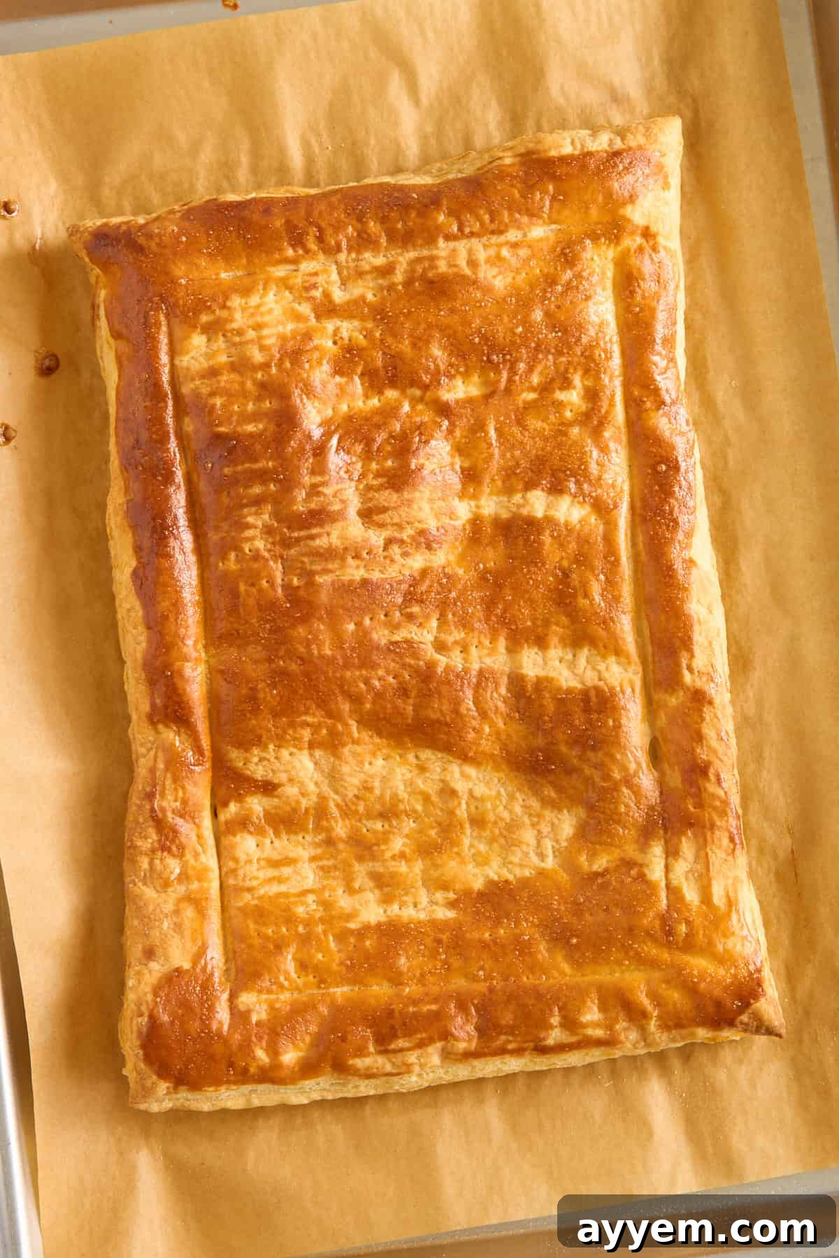 A rectangular golden-brown pastry with a shiny, flaky crust sits on a sheet of parchment paper. The edges are crimped, and the top shows light bake marks.