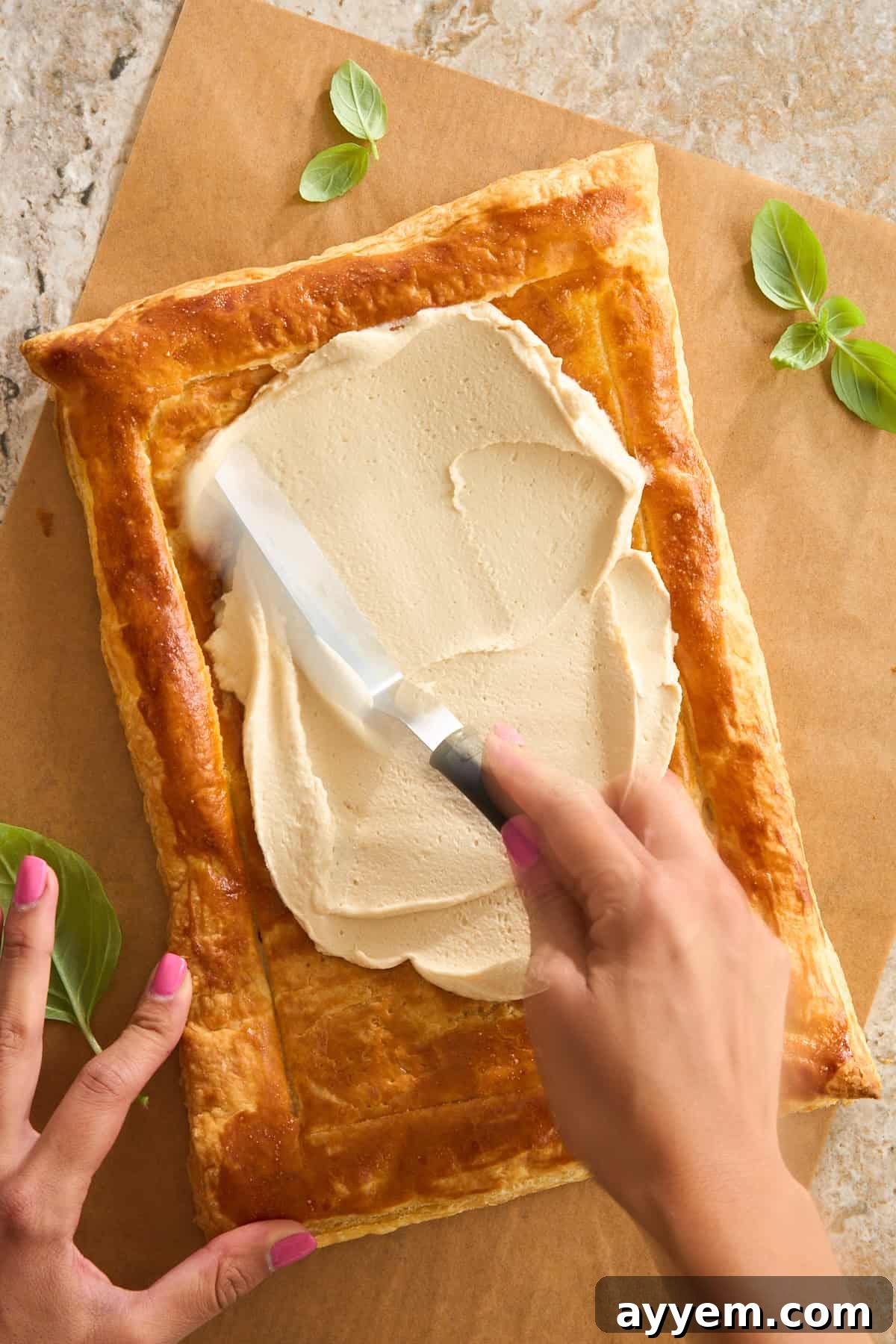 A person spreads a creamy, light-colored filling onto a rectangular, golden-brown puff pastry crust using an offset spatula. Fresh basil leaves and parchment paper are nearby.