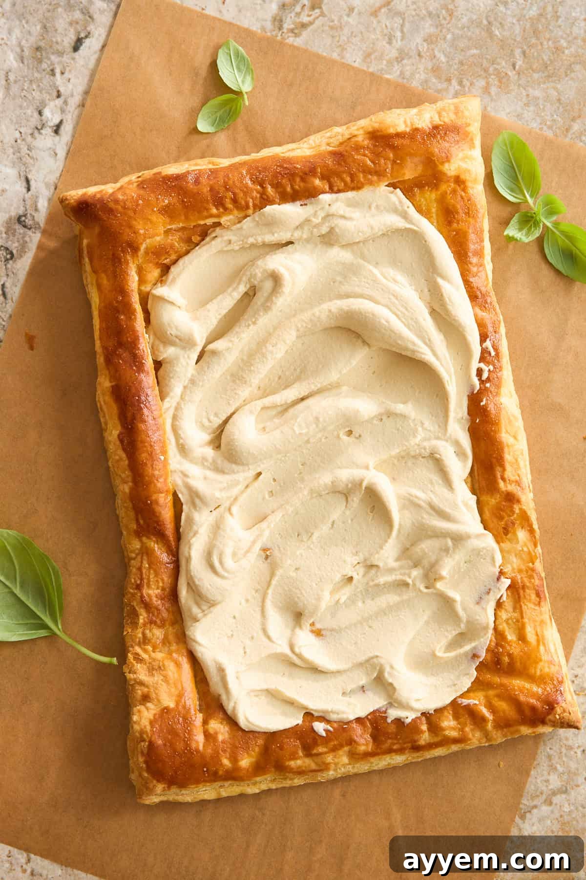 A rectangular sheet of golden-brown puff pastry with a thick layer of creamy spread on top, garnished with a few fresh green basil leaves on the side, on brown parchment paper.