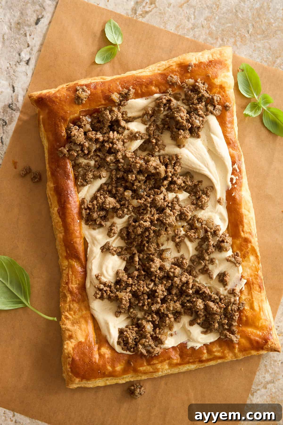 A rectangular puff pastry topped with a creamy spread and crumbled cooked sausage sits on brown parchment paper, garnished with fresh basil leaves on the side.