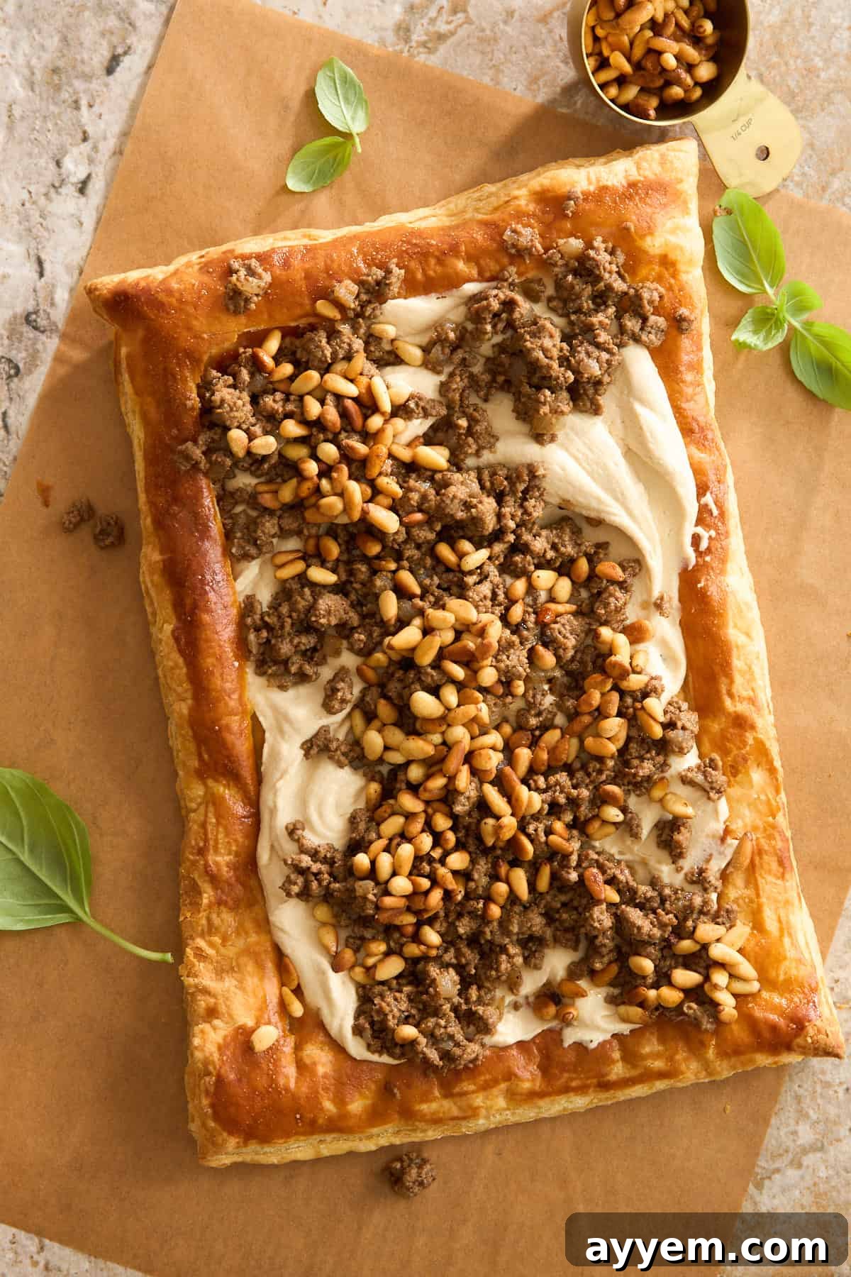A rectangular puff pastry tart topped with creamy spread, seasoned ground meat, and toasted pine nuts, garnished with fresh basil leaves on parchment paper.