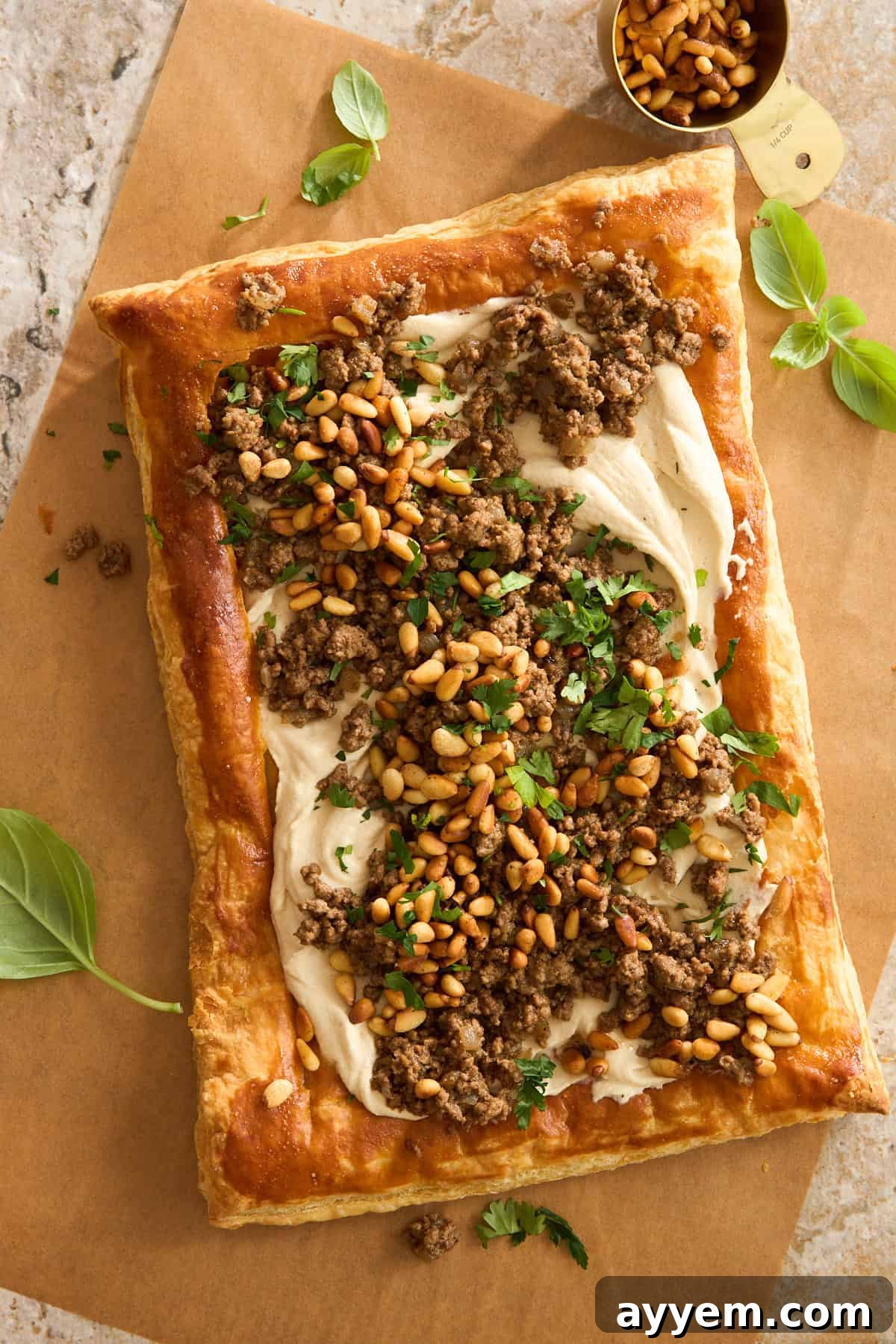 A rectangular puff pastry topped with creamy hummus, cooked ground beef, toasted pine nuts, and chopped parsley sits on parchment paper with basil leaves and a small cup of pine nuts nearby.