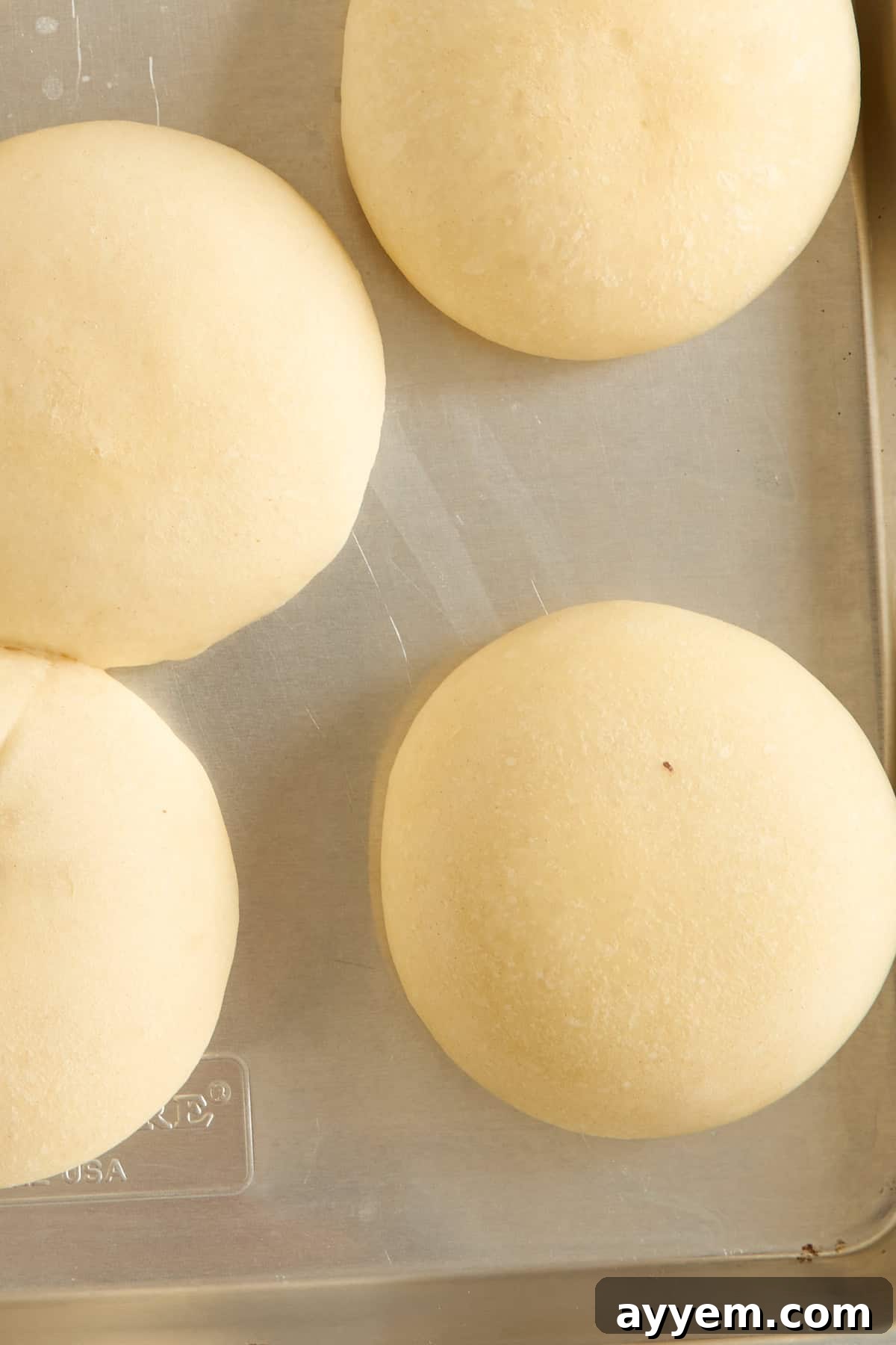 Four round, unbaked dough balls are arranged on a metal baking tray, ready to be baked. The surface of the dough is smooth and lightly dusted with flour.
