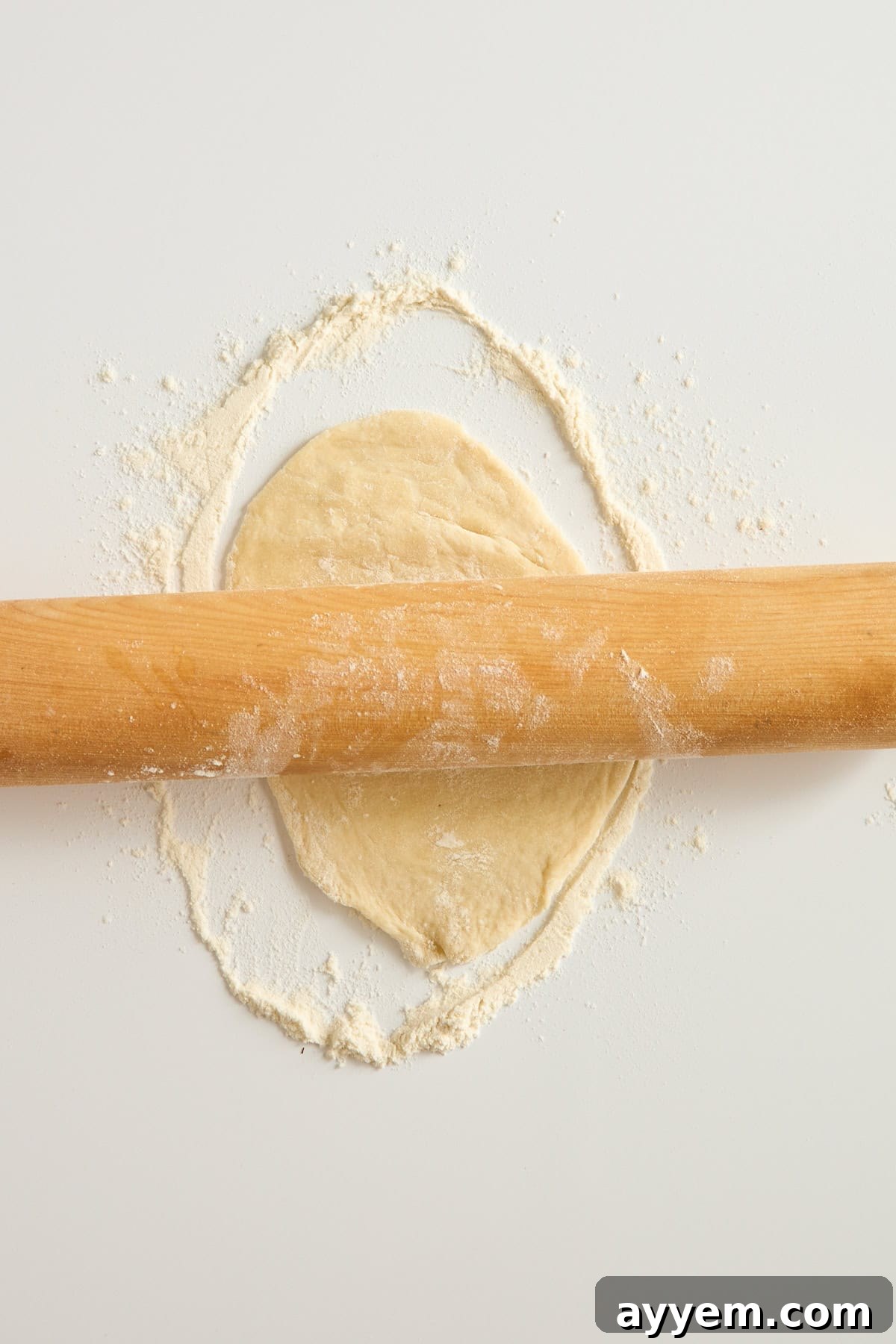 A wooden rolling pin rolls out a circle of dough on a white surface dusted with flour.