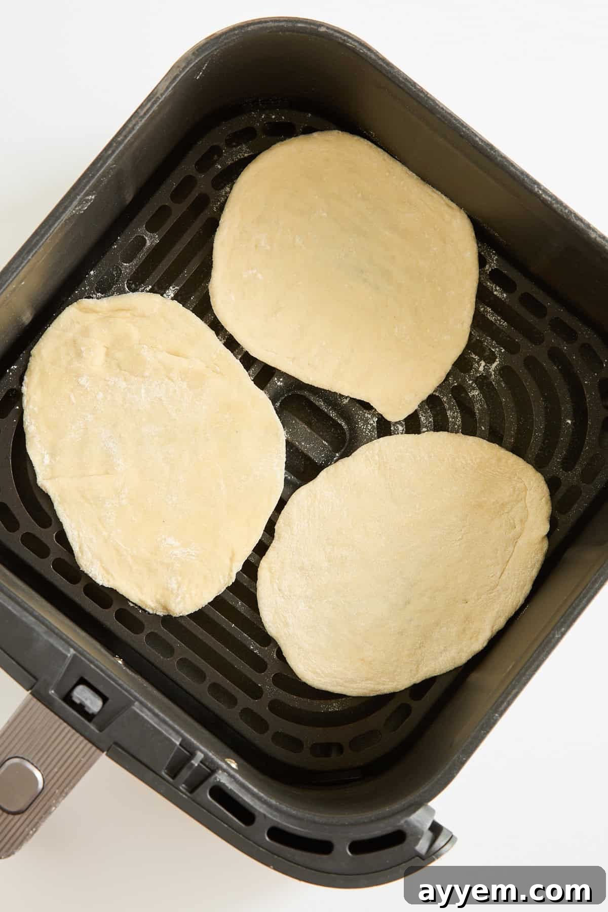 Three pieces of raw flatbread dough are placed inside the basket of an air fryer, ready to be cooked. The basket is black with a nonstick, perforated surface.