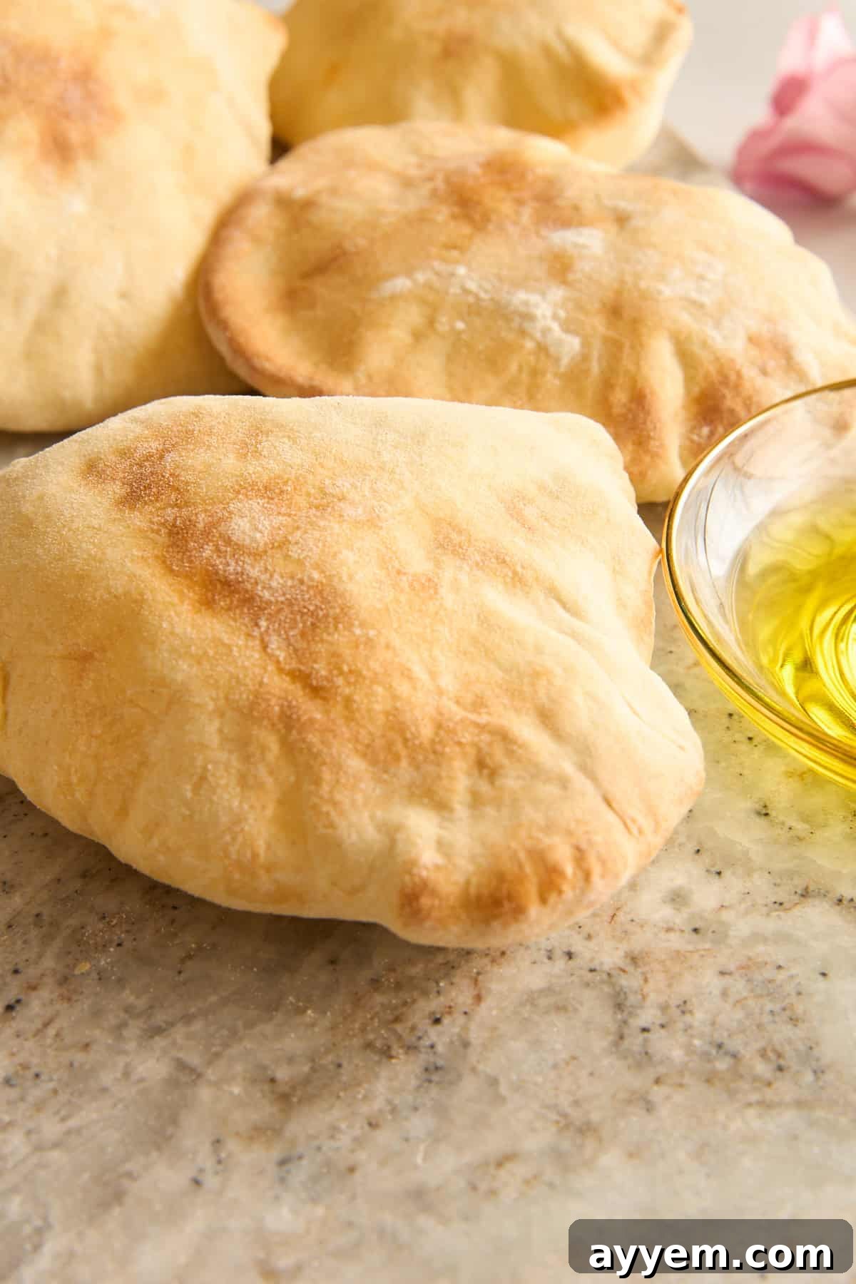 Three pieces of pita bread rest on a light-colored stone surface next to a small glass bowl filled with olive oil, ready for dipping.