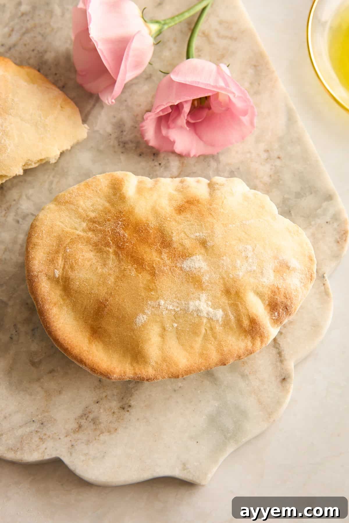 A golden-brown pita bread sits on a marble surface next to two pink flowers and a partial view of a glass dish with olive oil.