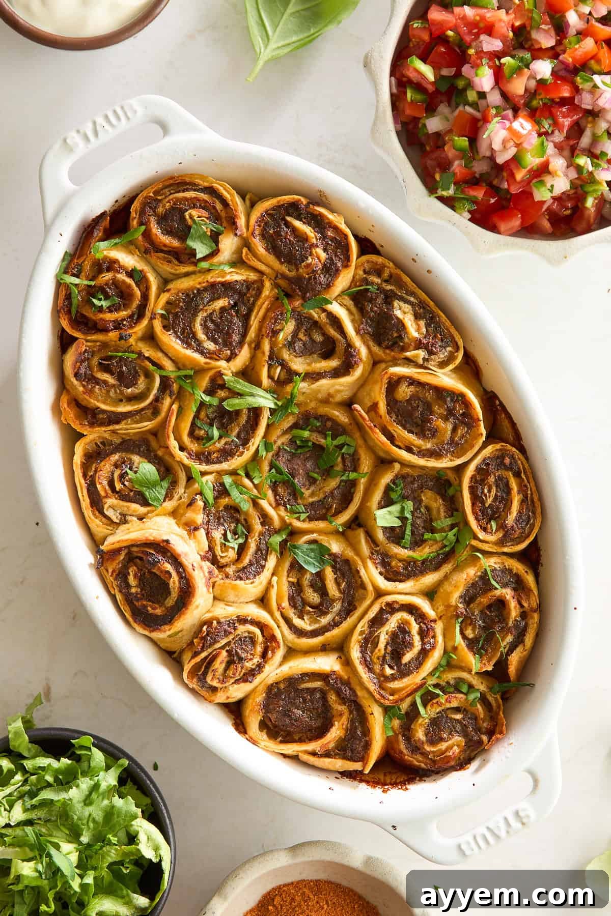 A white oval dish filled with golden-brown, spiral pinwheel rolls topped with chopped fresh herbs. Surrounding the dish are bowls of shredded lettuce, pico de gallo, sauce, and taco seasoning.