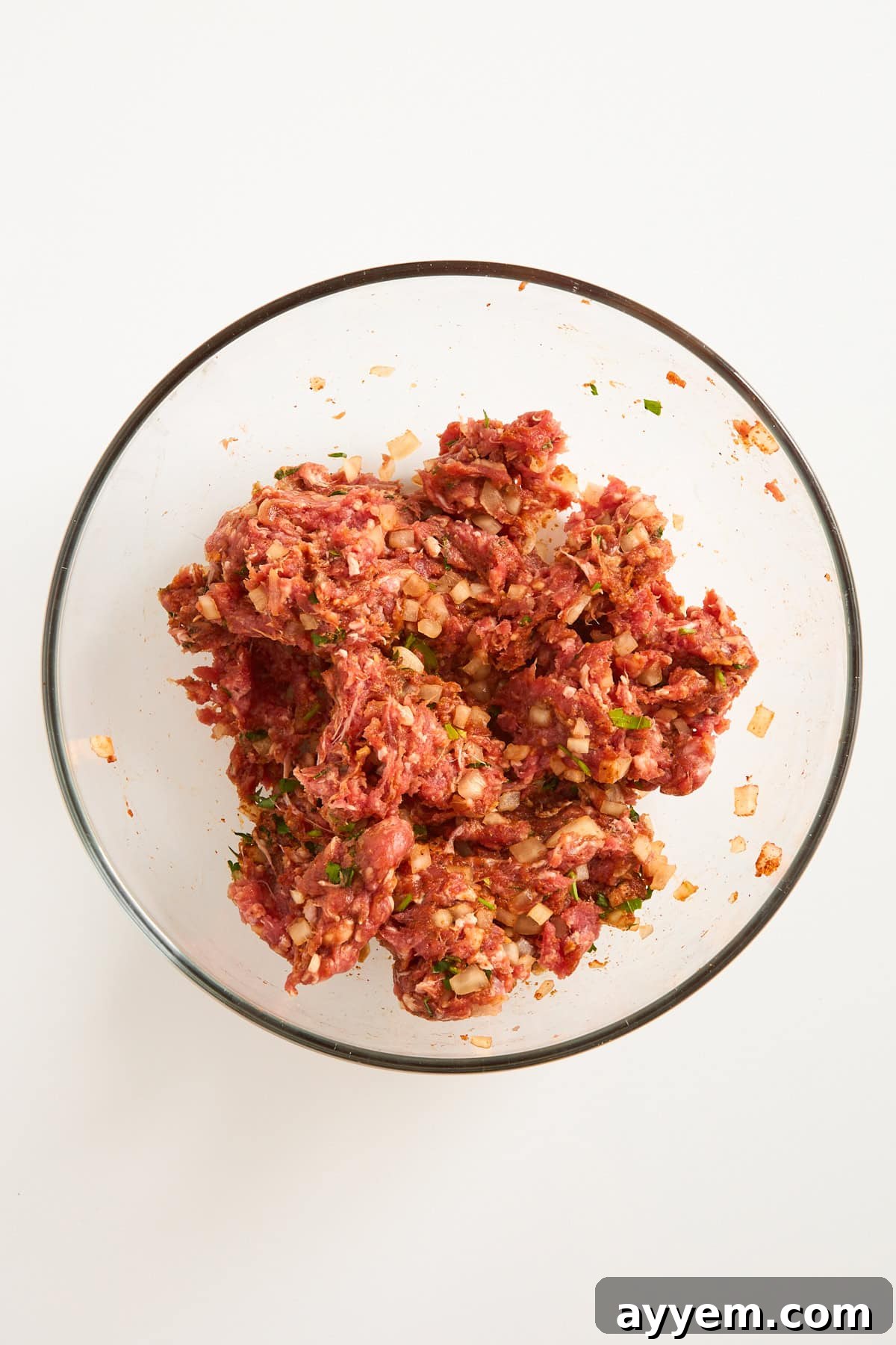 A clear glass bowl filled with a mixture of raw ground meat, chopped onions, herbs, and spices on a white background.
