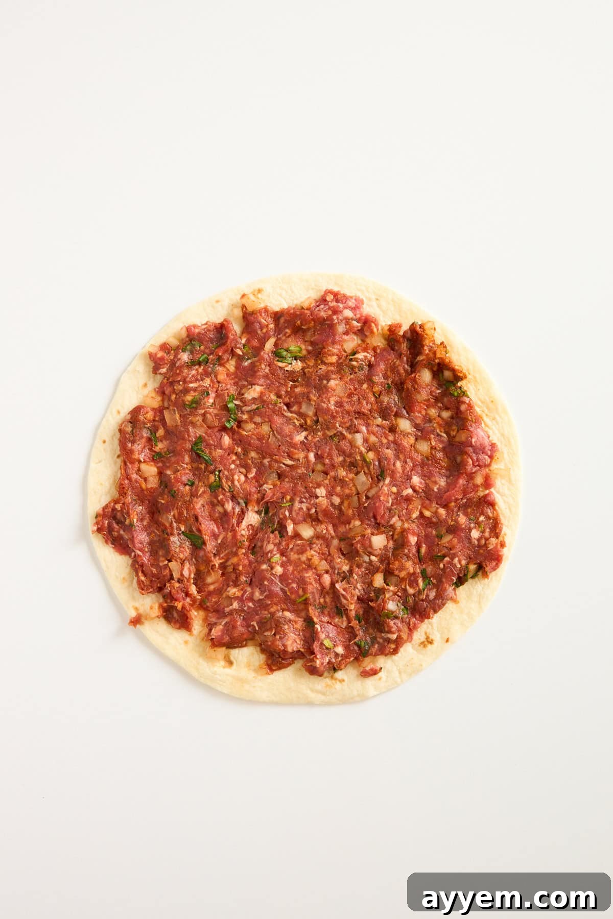 A round piece of flatbread topped with a raw, finely minced meat mixture, spread evenly to the edges, on a plain white background.
