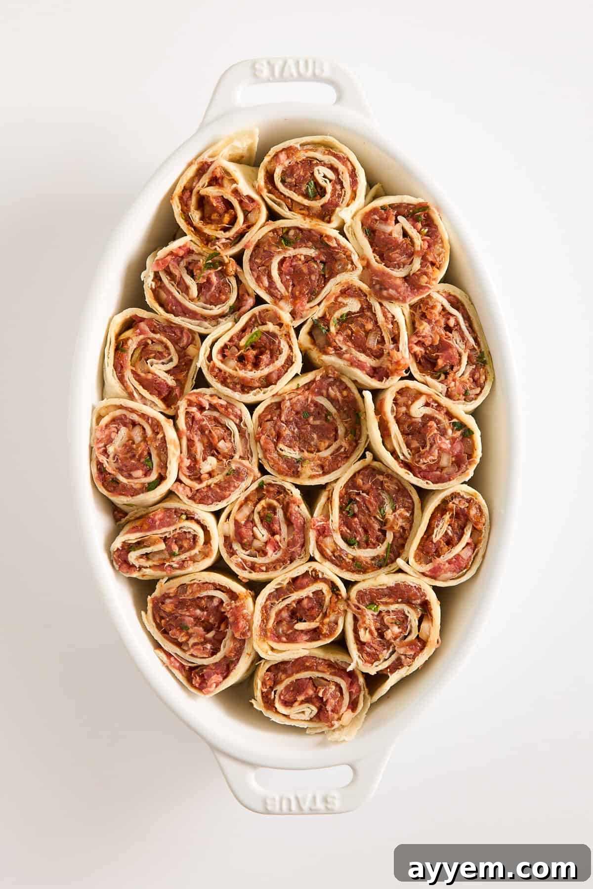 A white oval baking dish filled with neatly arranged, uncooked meat pinwheels made from rolled dough with a visible minced meat and herb filling. The dish is placed on a plain white surface.