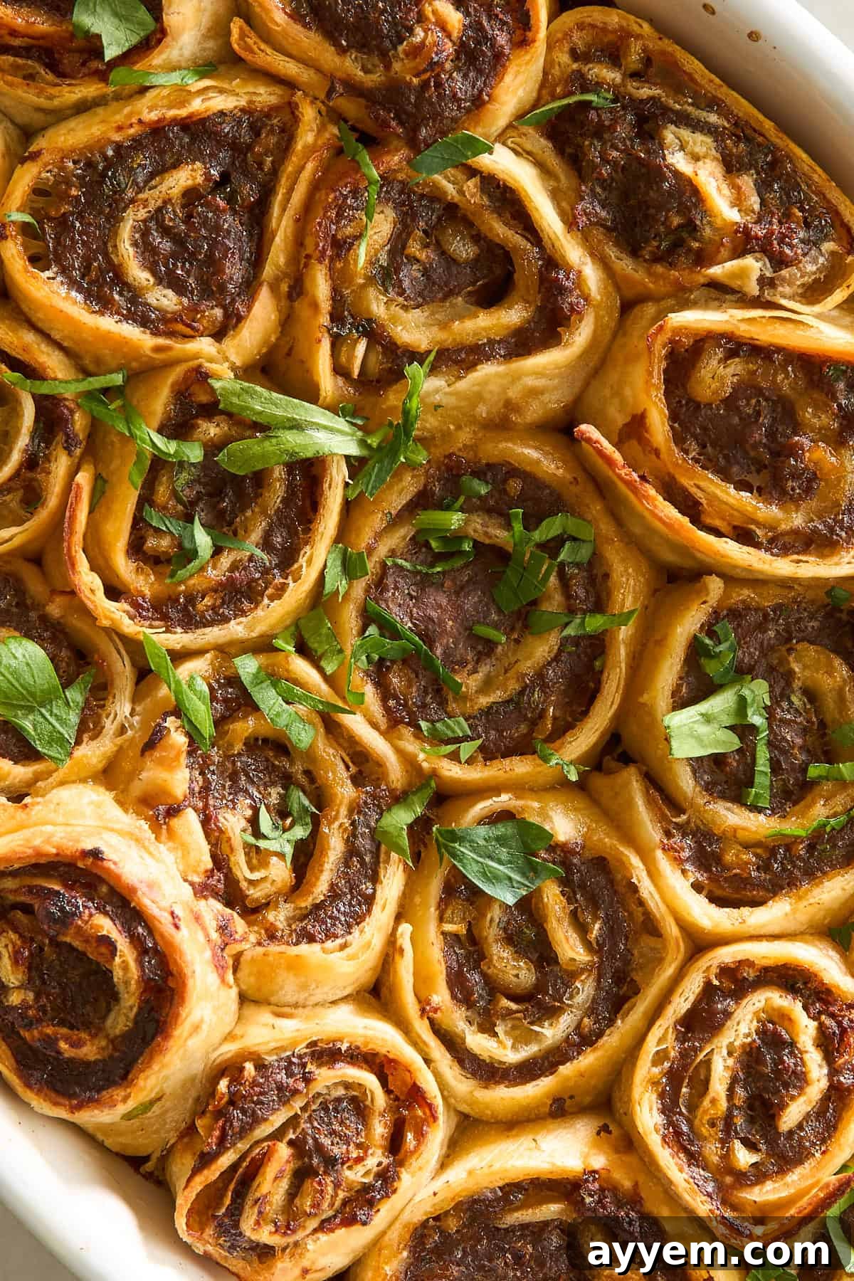 Golden-brown pinwheel pastries filled with a savory meat mixture, baked in a dish and garnished with fresh chopped parsley. The rolls are closely packed and have a crispy, flaky texture.