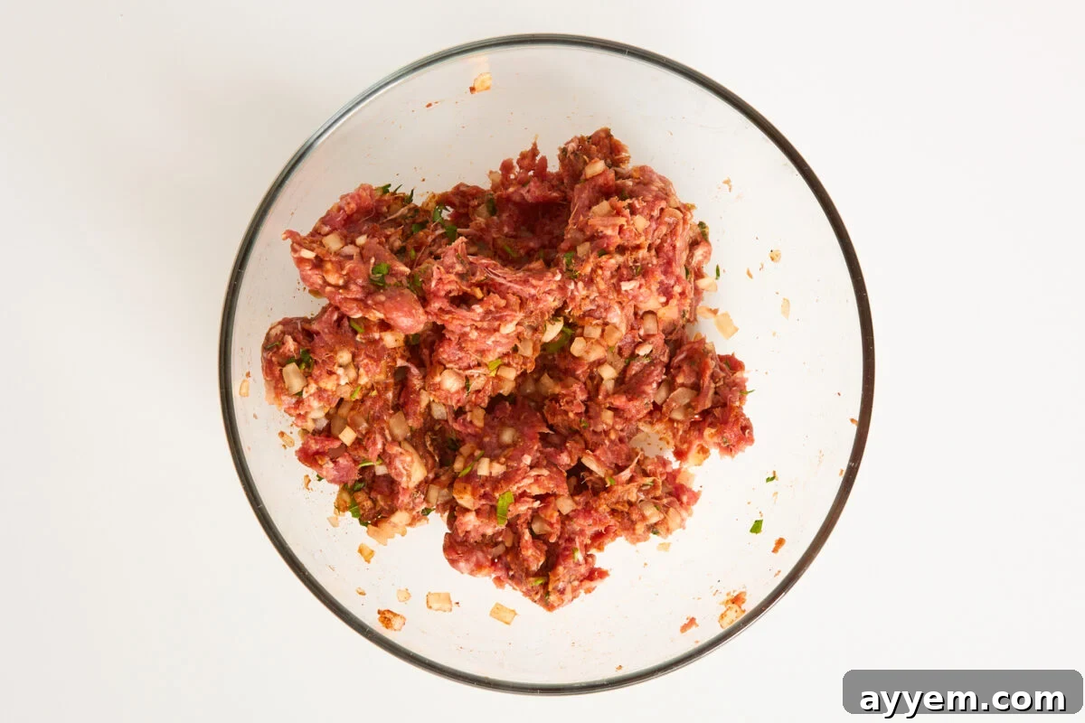A clear glass bowl filled with a mixture of raw ground meat, chopped onions, herbs, and spices on a white background.