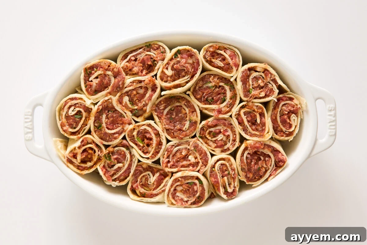 A white oval baking dish filled with neatly arranged, uncooked meat pinwheels made from rolled dough with a visible minced meat and herb filling. The dish is placed on a plain white surface.