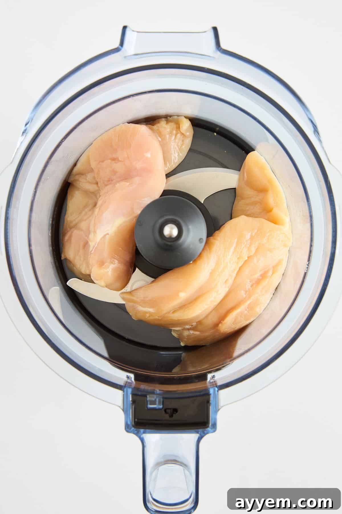 Raw chicken breast pieces placed inside a food processor, viewed from above, ready to be processed. The food processor has a clear plastic bowl and a sharp metal blade in the center.