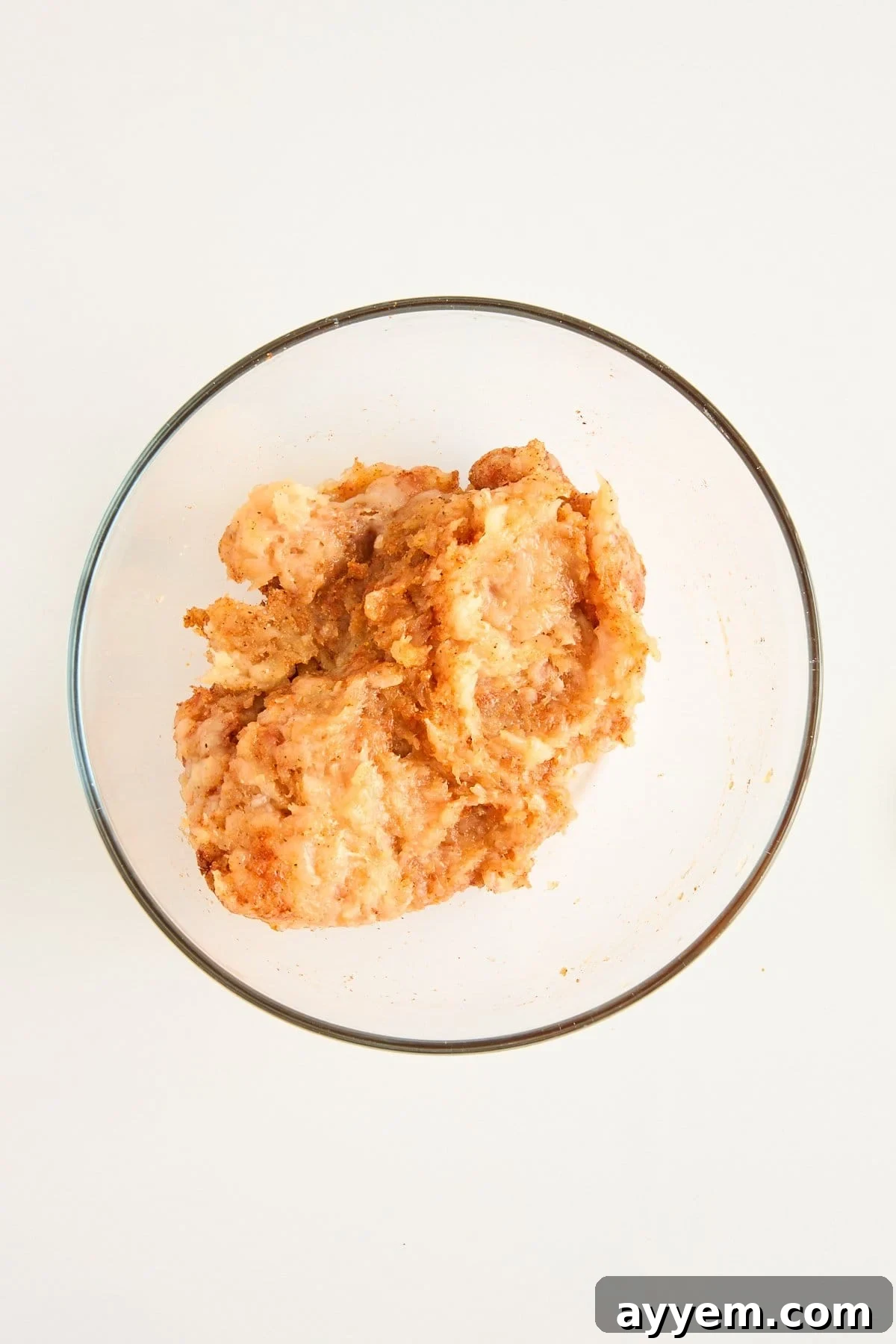 A clear glass bowl containing a mixture of ground meat and seasonings on a white surface.