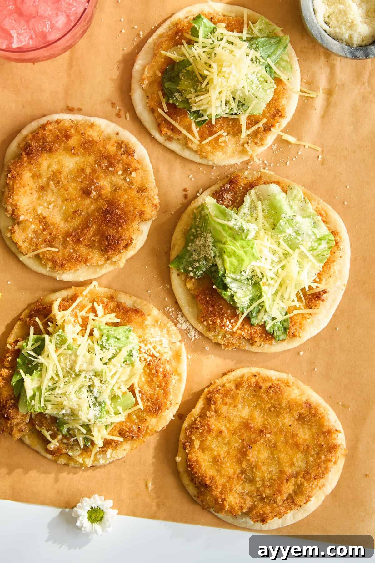 Five small round flatbreads are topped with breaded chicken, lettuce, shredded cheese, and grated parmesan, arranged on parchment paper with a small bowl of parmesan in the corner.