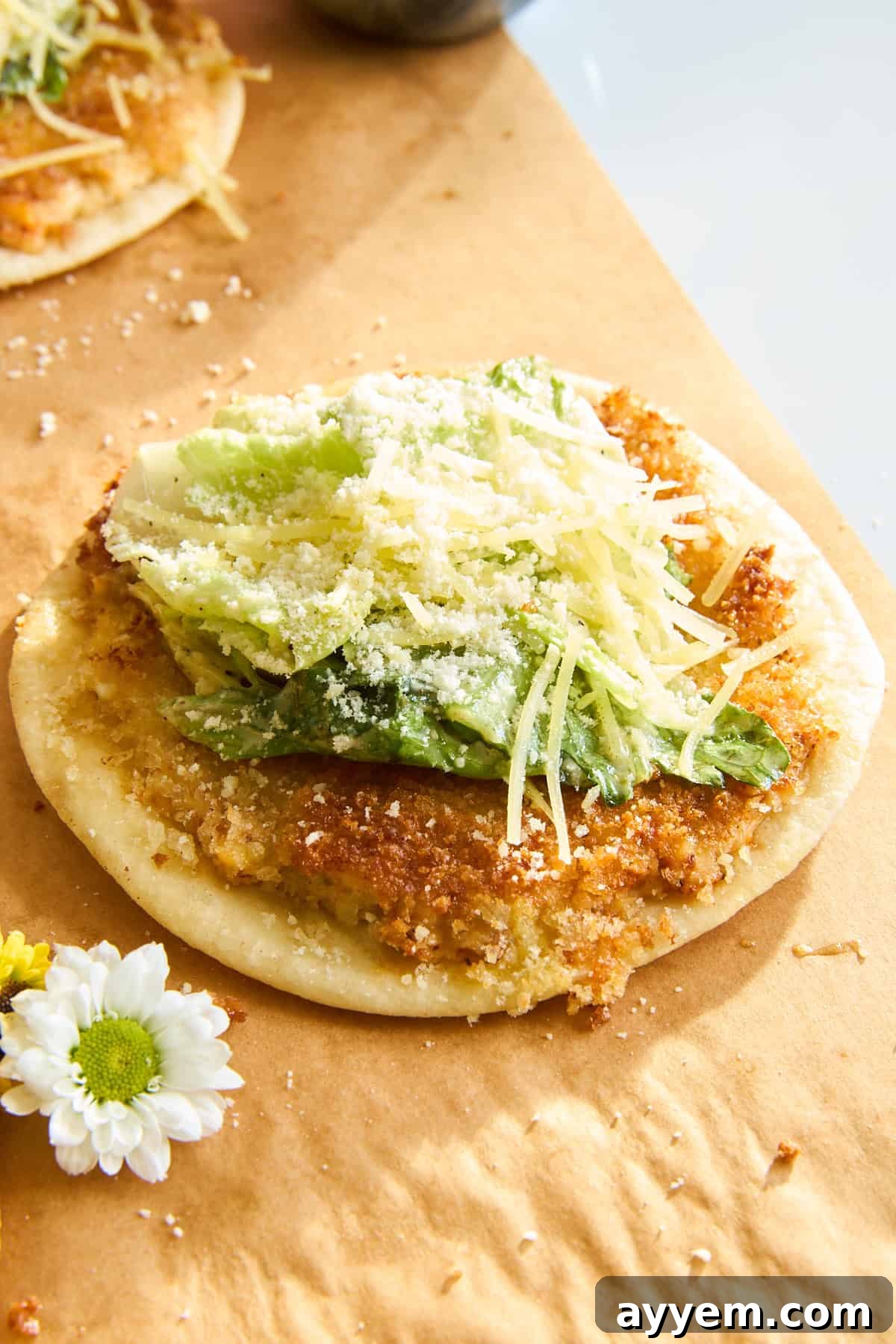 A crispy round flatbread topped with fresh green lettuce, grated cheese, and a sprinkle of parmesan, served on brown parchment paper with a white daisy flower beside it.