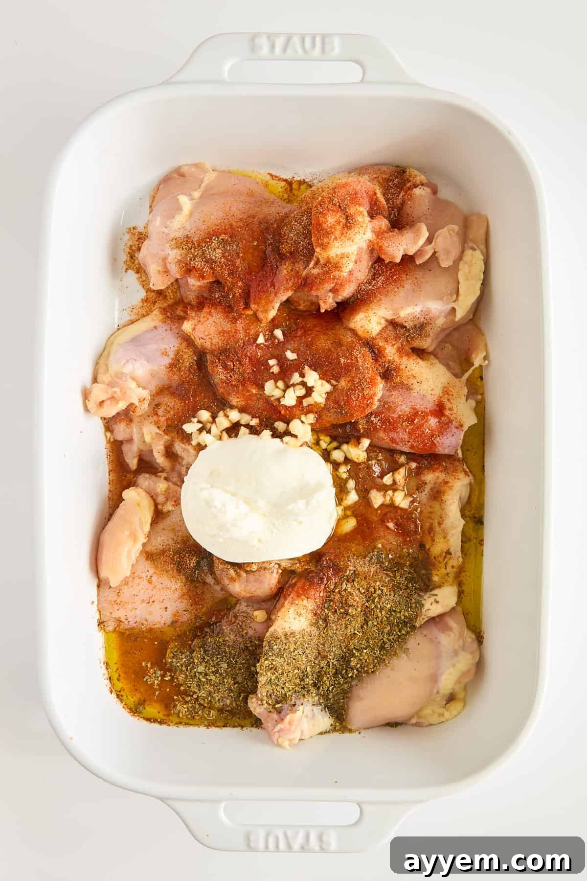 Raw chicken thighs in a white baking dish, topped with minced garlic, herbs, paprika, ground spices, and a dollop of sour cream or yogurt, ready to be mixed or marinated.