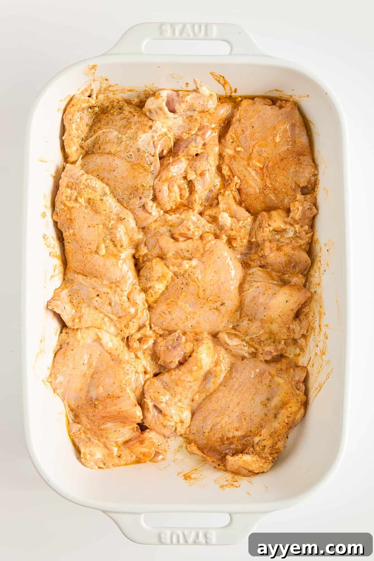 Raw chicken pieces coated in a reddish-orange marinade are arranged in a single layer in a white rectangular baking dish. The chicken is uncooked and evenly distributed in the dish, ready for marination.