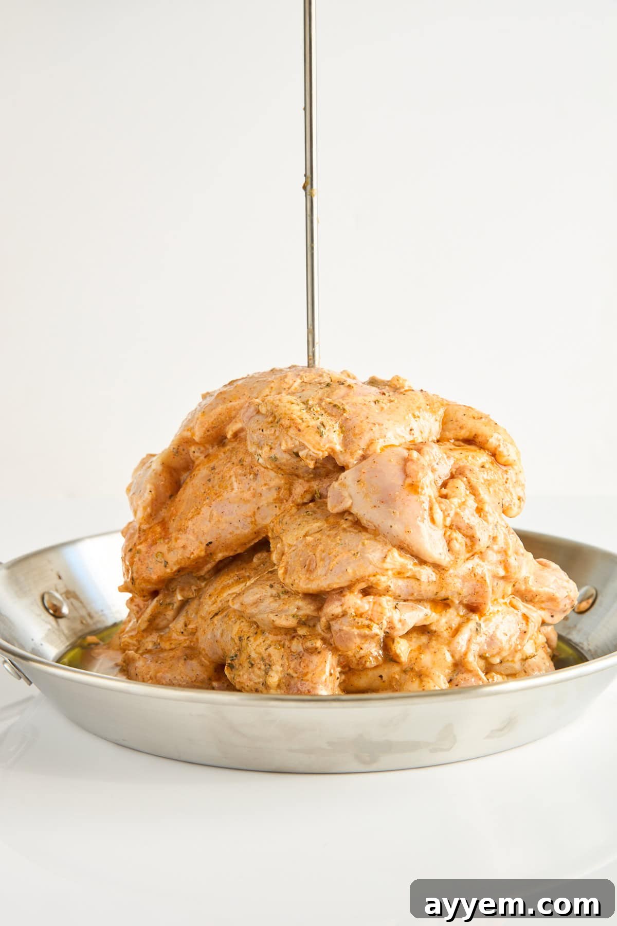 A stack of marinated raw chicken pieces is piled on a vertical skewer in a shallow metal pan, ready to be cooked, against a plain white background.