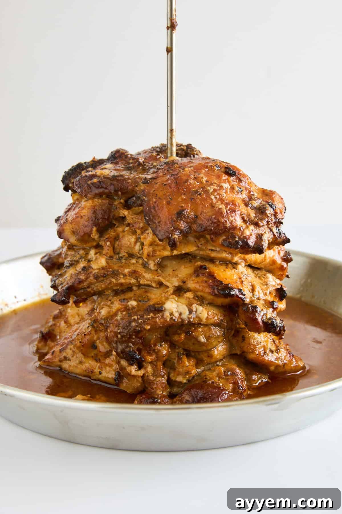 A stack of seasoned, roasted meat slices skewered vertically on a metal rod, sitting on a round metal tray with juices collecting at the bottom, mimicking a traditional gyro spit.