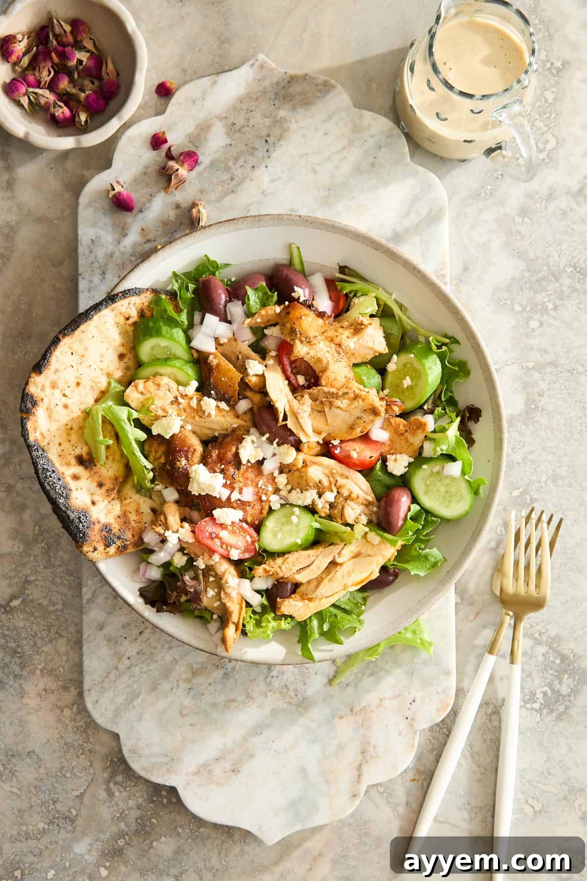 A bowl of fresh Greek salad with grilled chicken, crisp cucumber, cherry tomatoes, Kalamata olives, red onions, crumbled feta cheese, and mixed greens. A side of warm pita bread, a small jar of dressing, and cutlery are arranged on a marble surface, creating an inviting presentation.