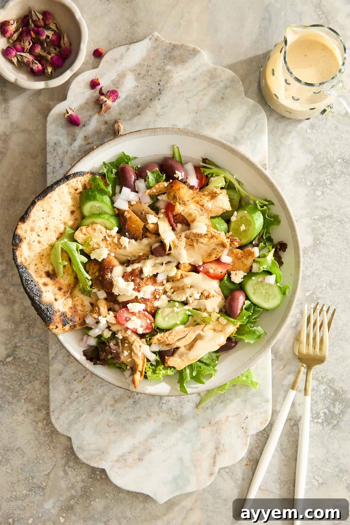 A bowl of fresh Greek salad with grilled chicken, pita bread, cucumbers, tomatoes, feta cheese, and olives on a marble surface, with a fork, knife, a small pitcher of dressing, and a bowl of dried rose petals nearby.