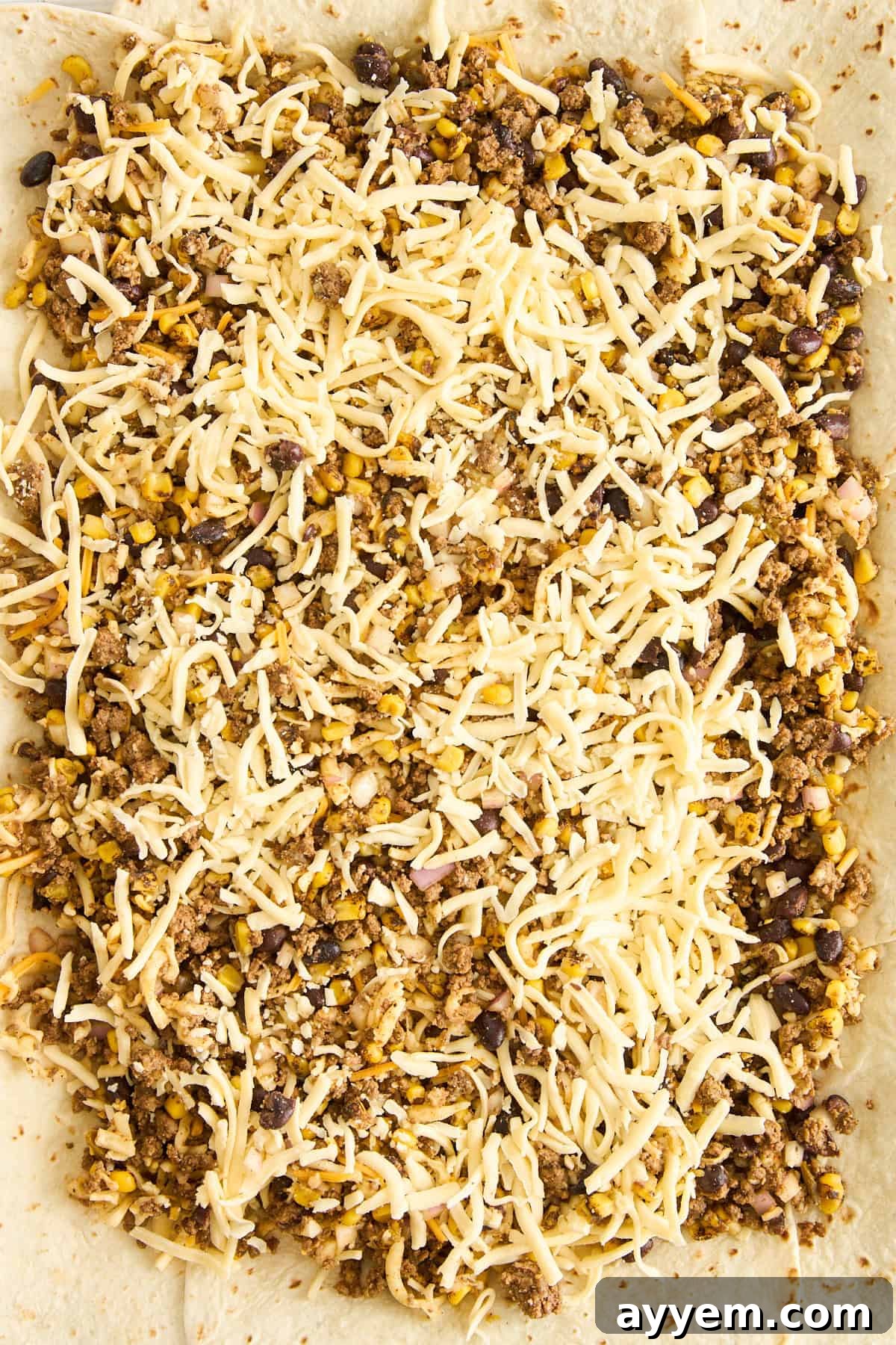 A close-up view of a flour tortilla topped with shredded cheese, cooked ground meat, black beans, corn, and rice, ready to be rolled or folded for a burrito or wrap.