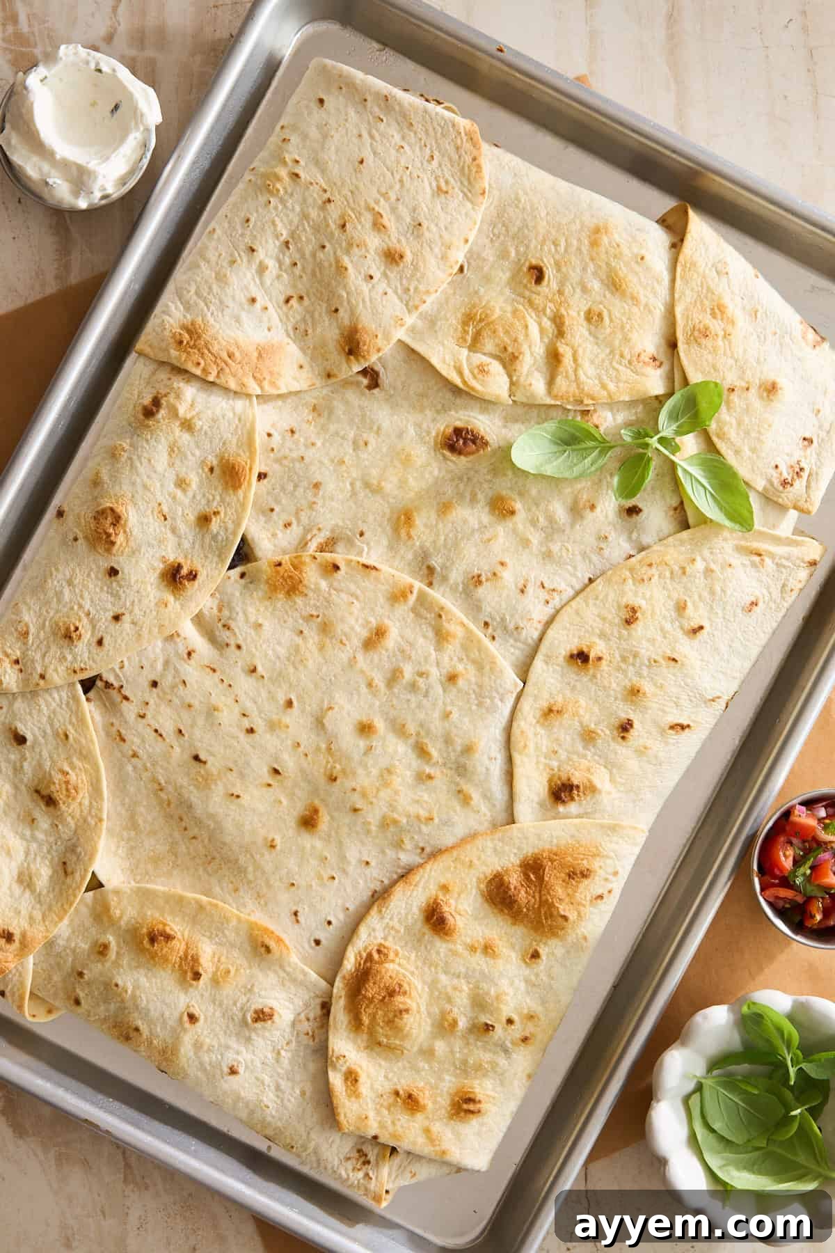 A baking sheet covered with overlapping toasted flour tortillas, garnished with fresh basil. Small bowls of sour cream and salsa sit nearby on a beige surface.