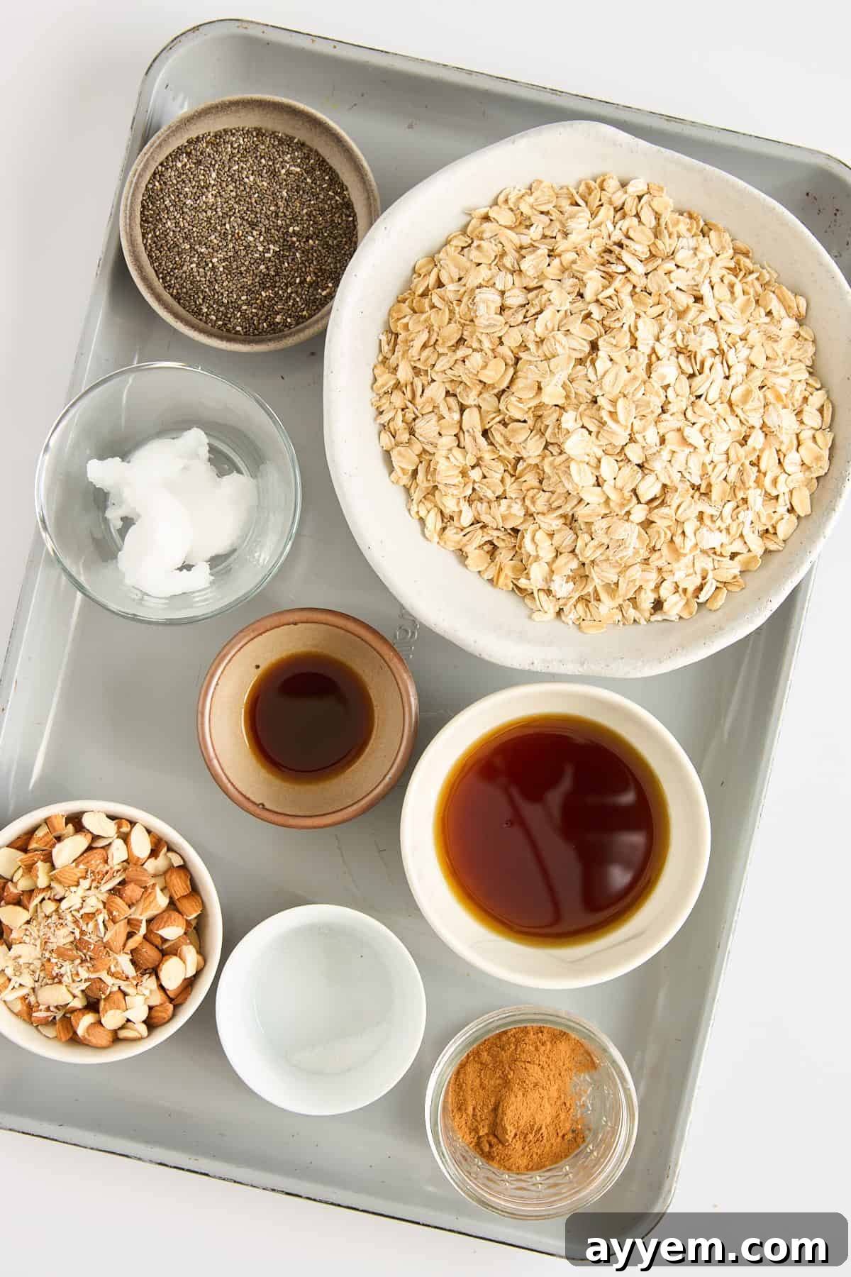 Crunchy Air Fryer Granola 3 A tray with bowls containing rolled oats, chia seeds, coconut oil, chopped almonds, vanilla extract, maple syrup, salt, and ground cinnamon arranged neatly for baking.