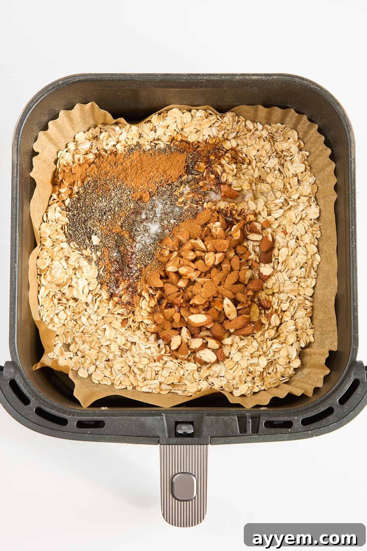 Crunchy Air Fryer Granola 4 An air fryer basket lined with parchment paper contains dry oats, chopped nuts, chia seeds, cinnamon, and a drizzle of honey, ready to be mixed.