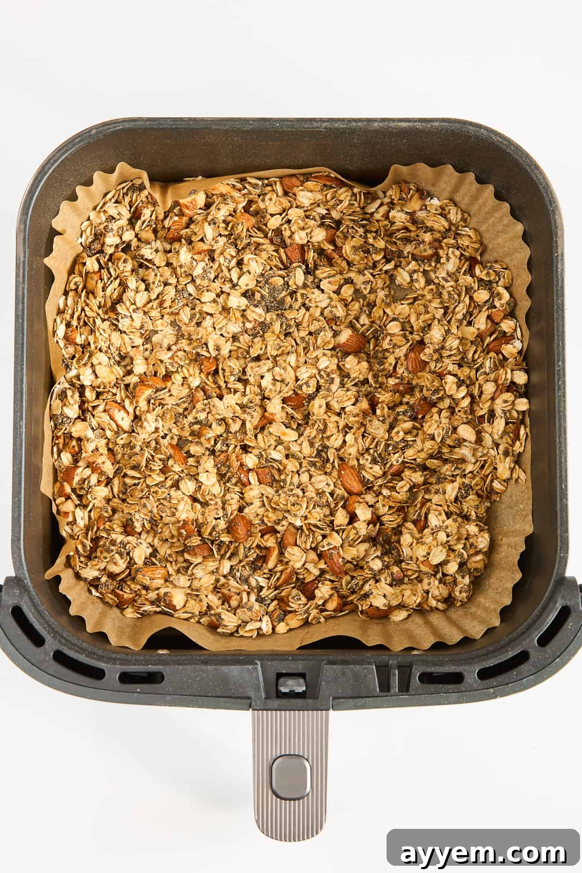 Crunchy Air Fryer Granola 5 A square air fryer basket lined with parchment paper contains an even layer of granola with visible oats and almonds, ready for baking.