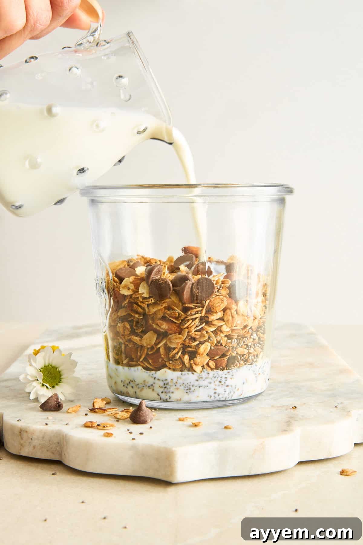 A hand pours milk from a glass pitcher into a clear jar filled with granola, chocolate chips, and chia seeds, sitting on a marble board with a small white flower nearby.