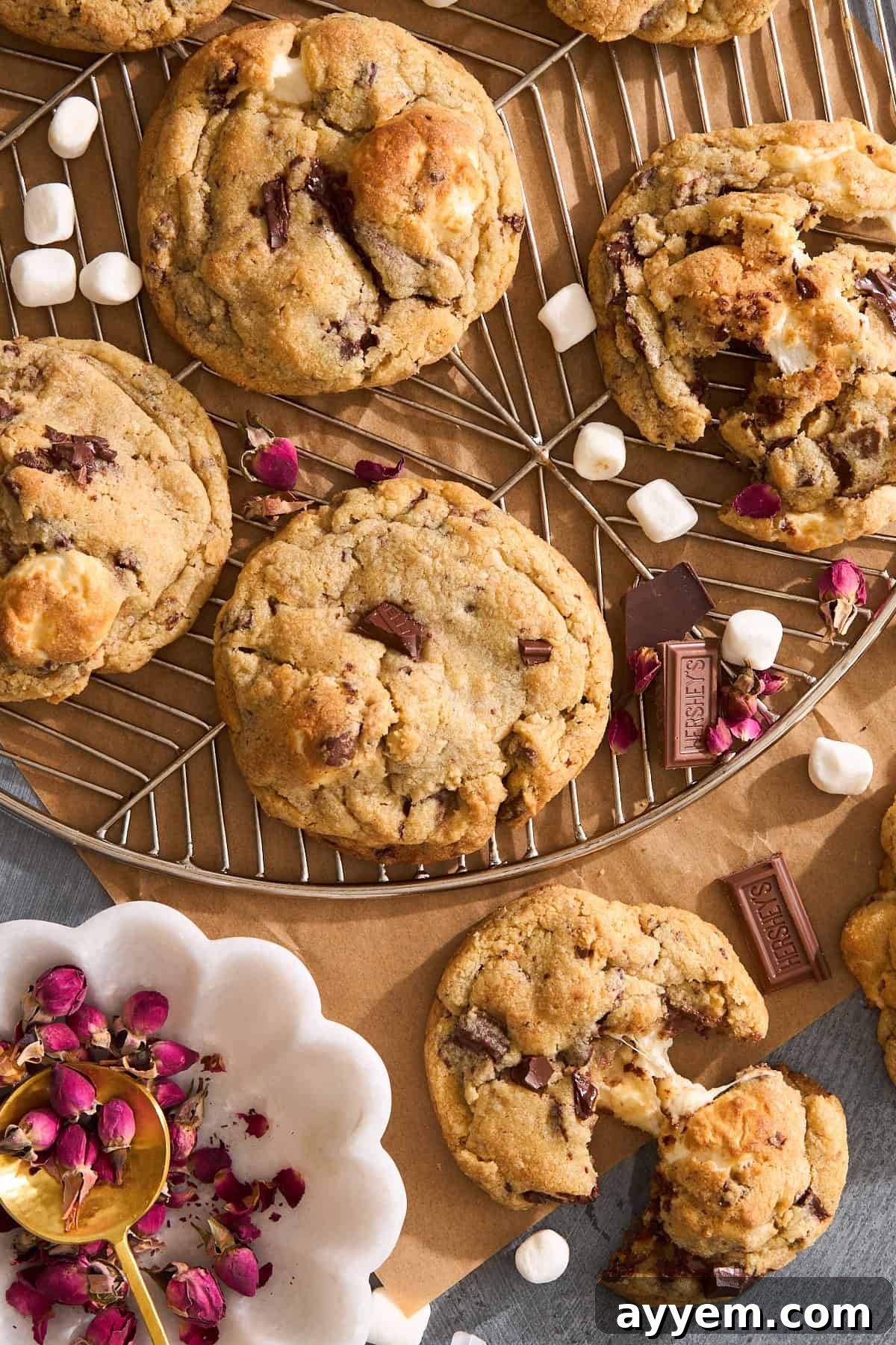 Why Are My Cookies So Flat 2 A batch of chocolate chip cookies with chunks of chocolate and marshmallows, some whole and some broken open, are displayed on a wire rack with small marshmallows and rose petals scattered around.